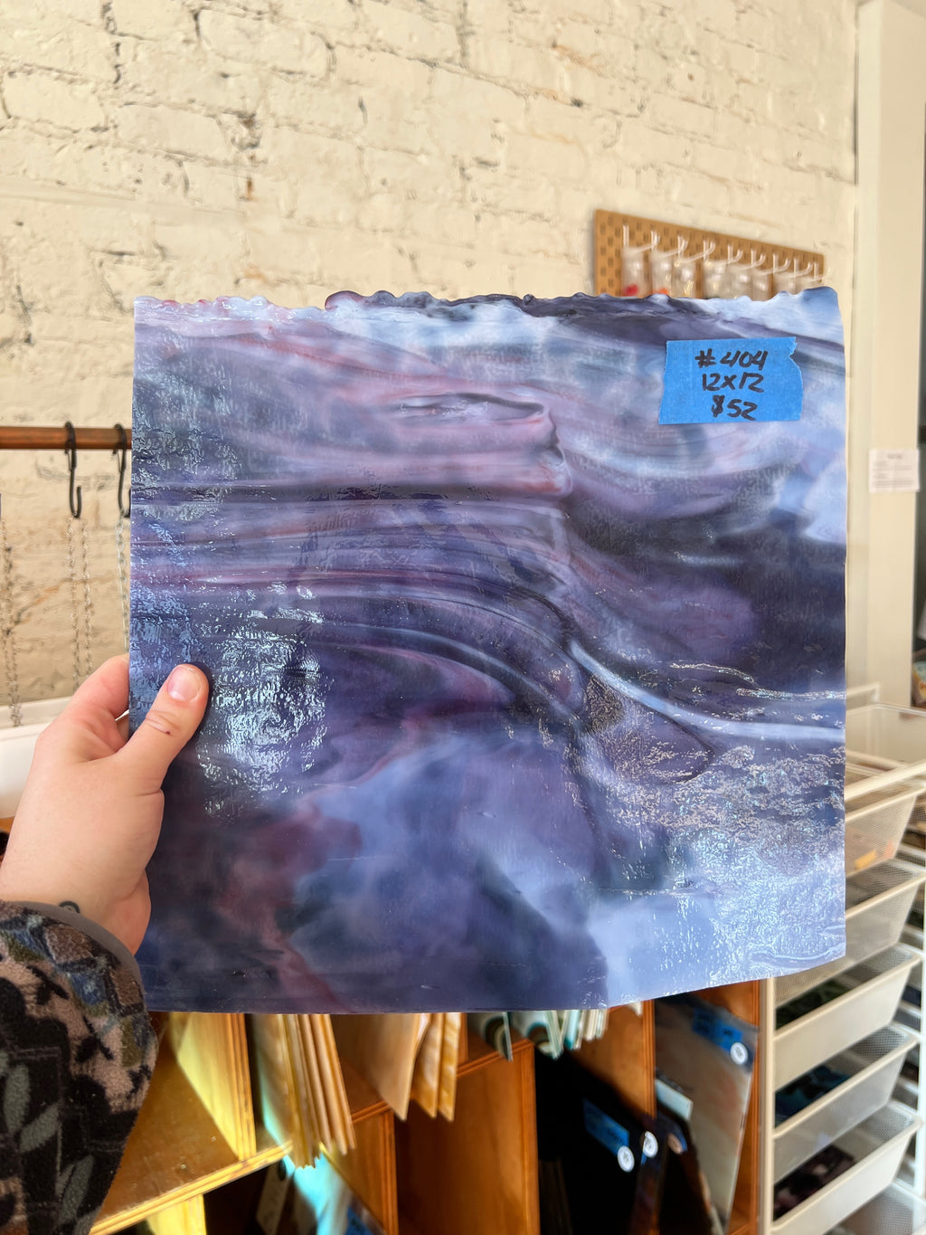 Hand holding a piece of abstract sheet glass with swirling purple and blue patterns against a brick wall.