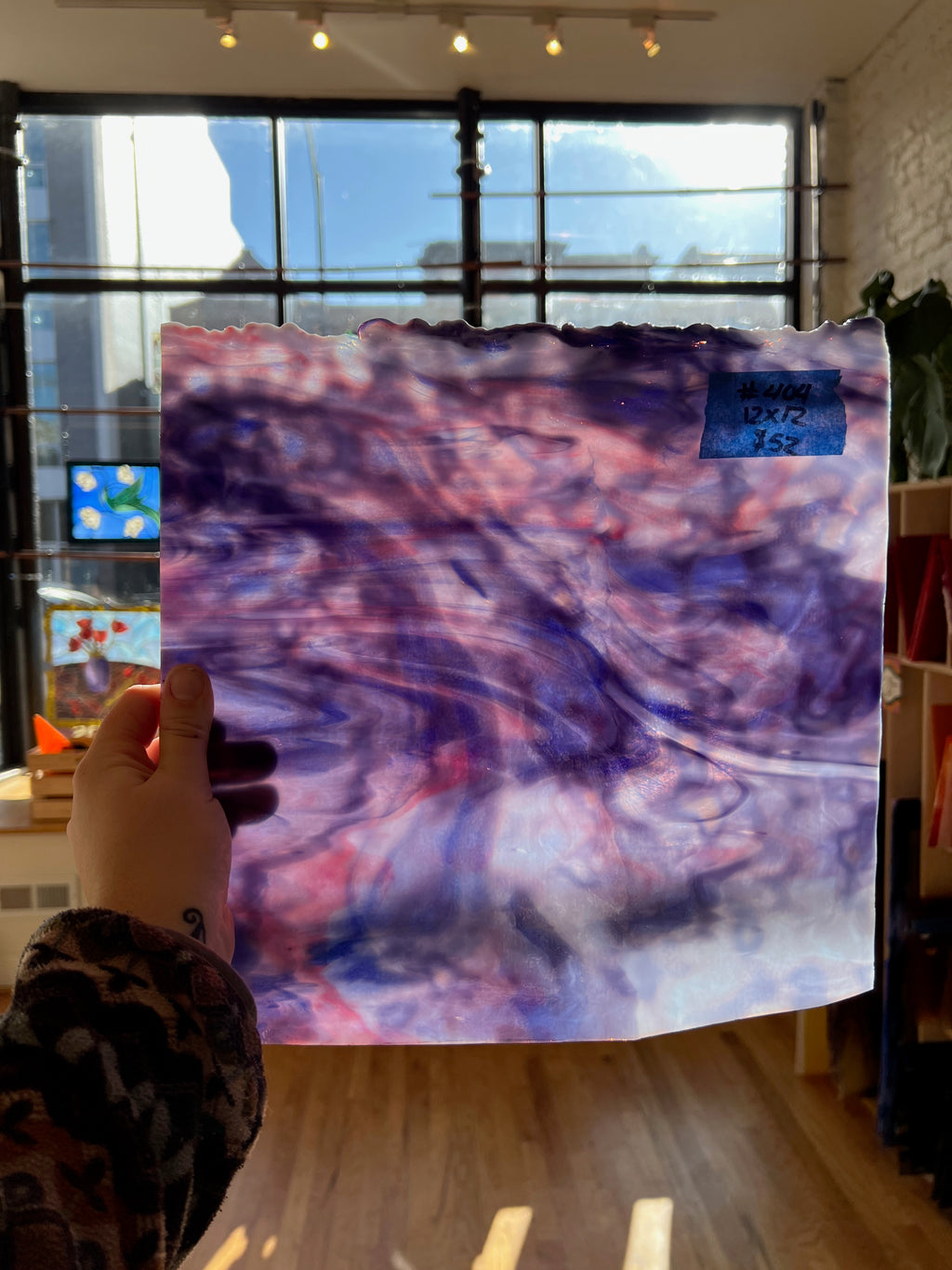 Hand holding a piece of sheet glass with abstract purple and pink design in an indoor setting.