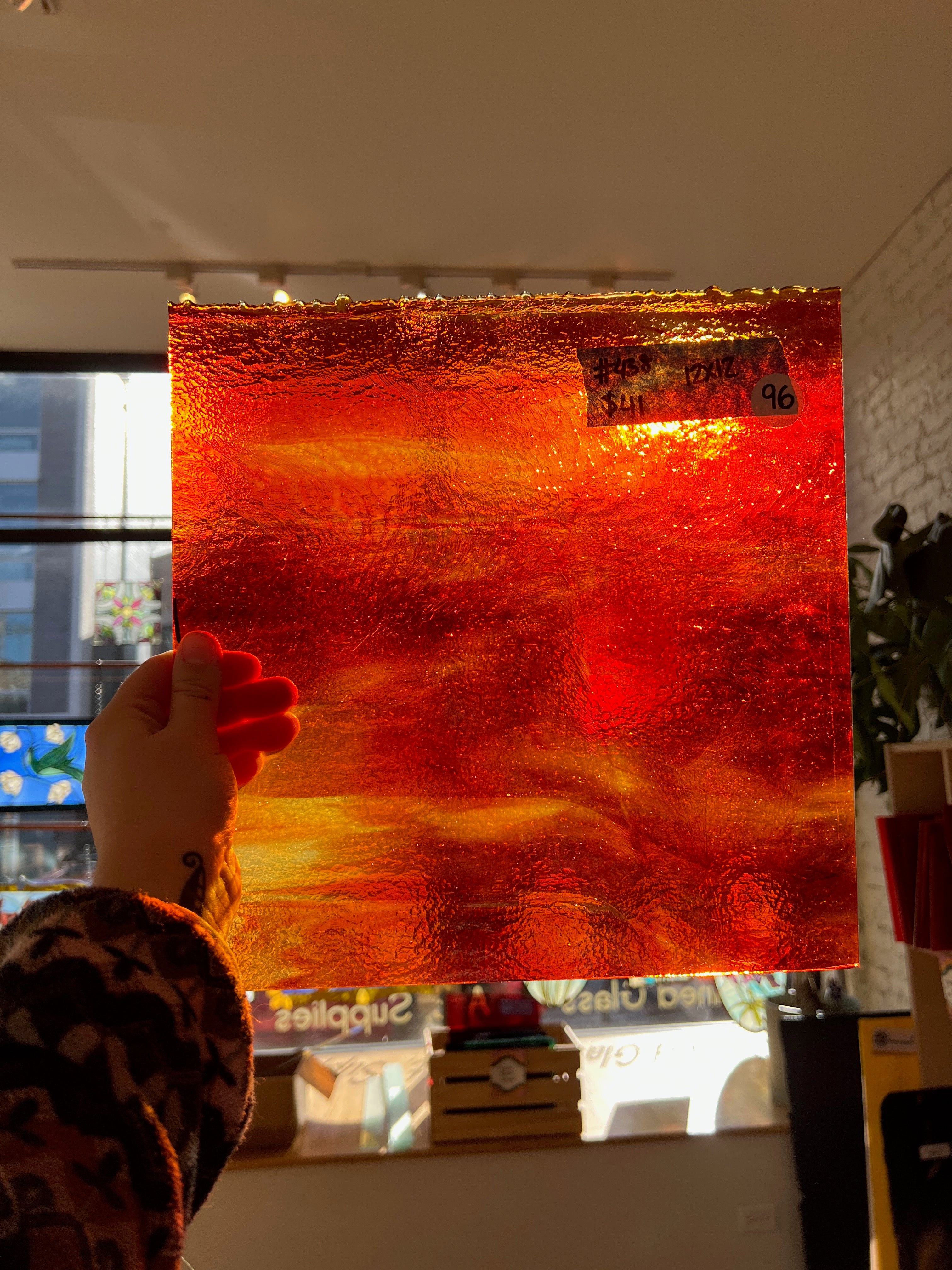 Hand holding a rectangular piece of orange and red textured glass sheet in front of a window.