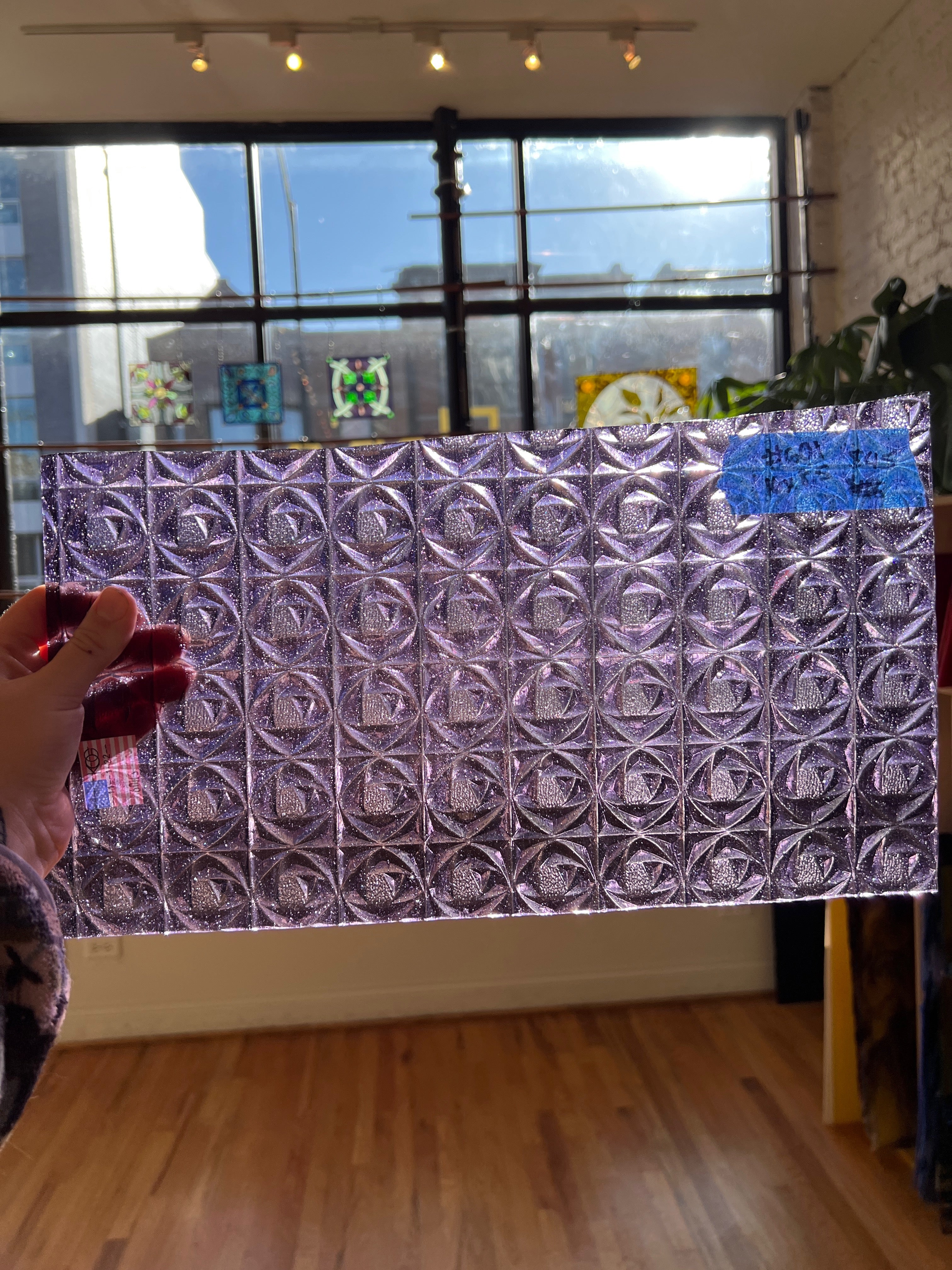 Person holding a rectangular metallic glass panel with a geometric pattern in a room with large windows.