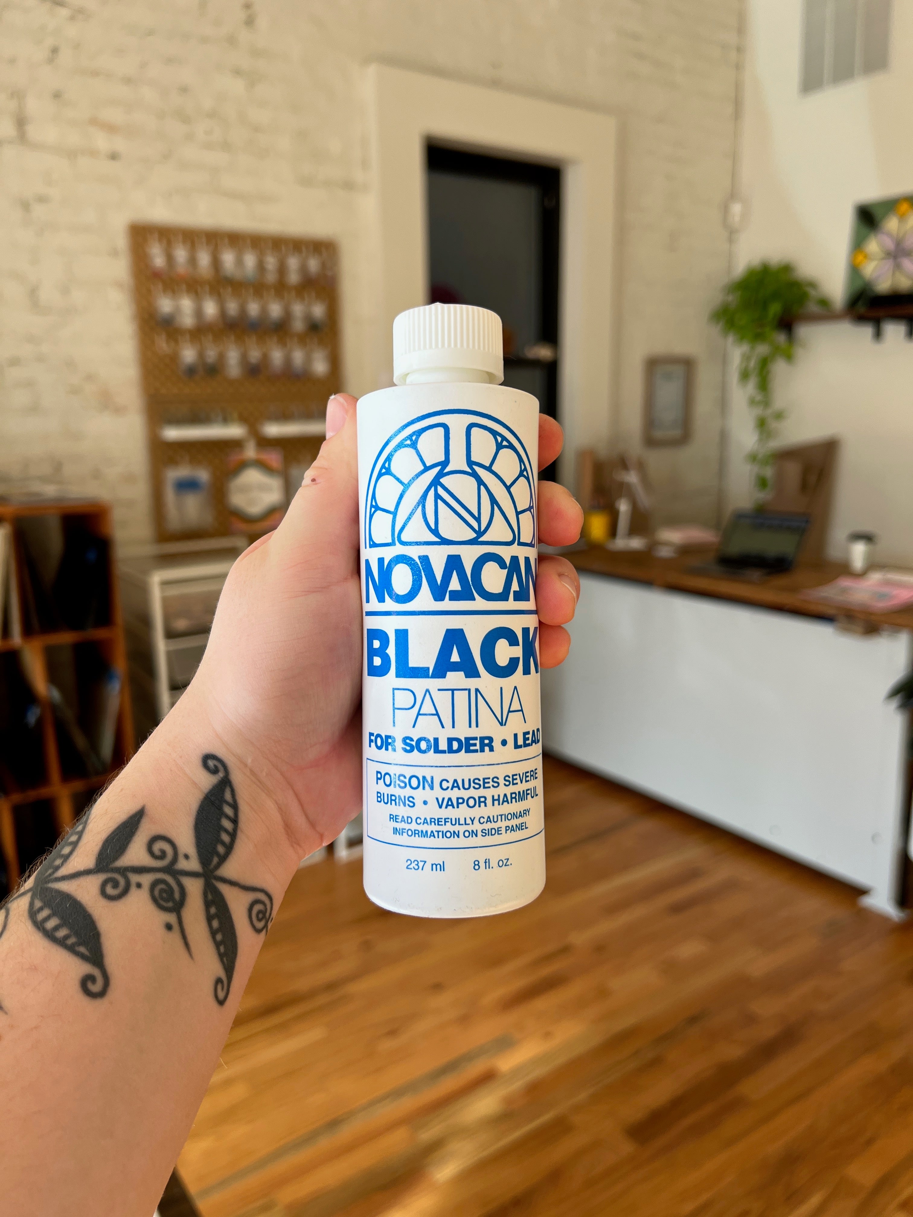 Hand holding a bottle of Nova Black Patina in a room with wooden floors and a white brick wall.