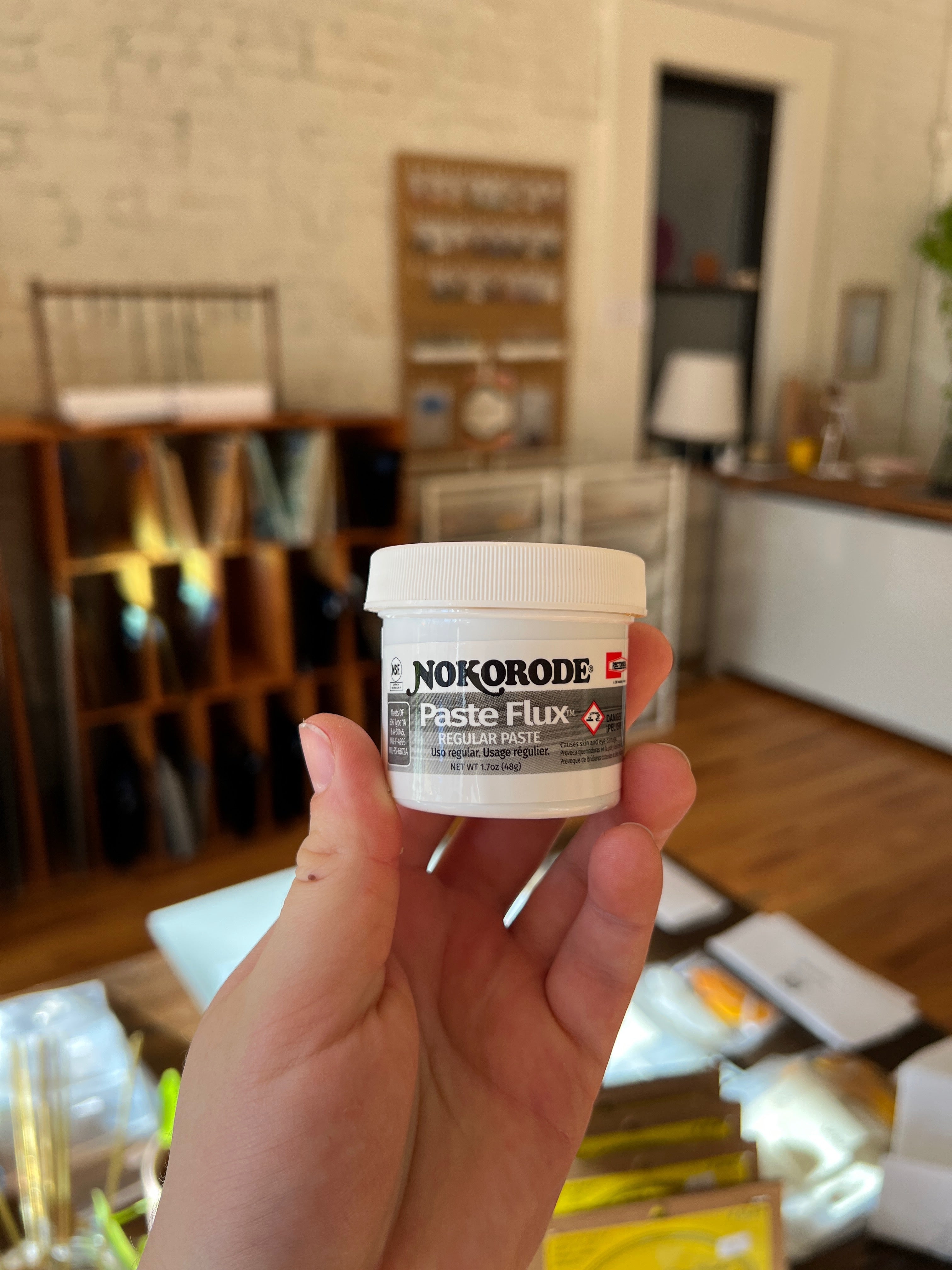 Hand holding a small container of Nokorode Paste Flux in an indoor setting.