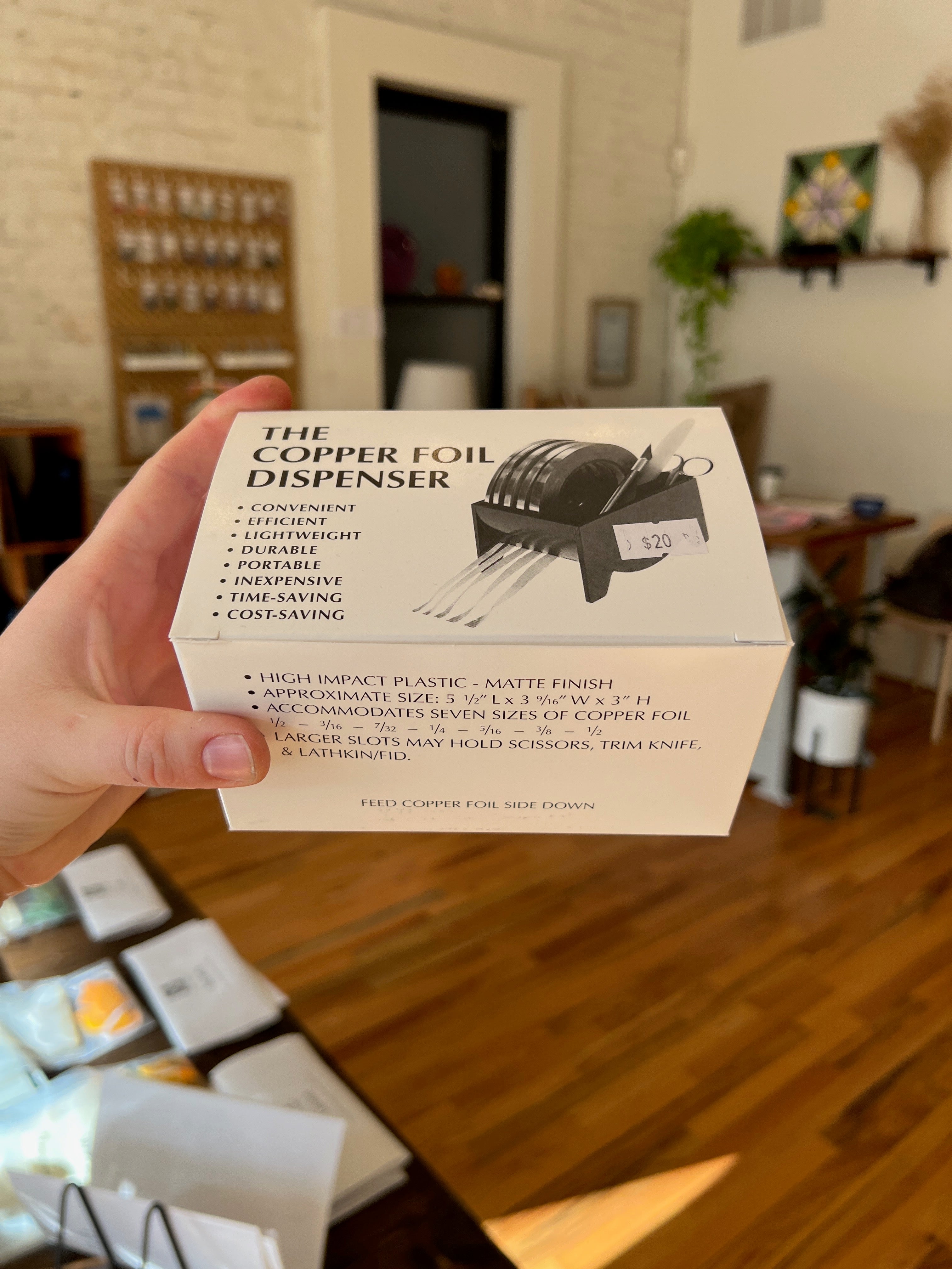Hand holding a box of 'The Copper Foil Dispenser' with a blurred indoor background.