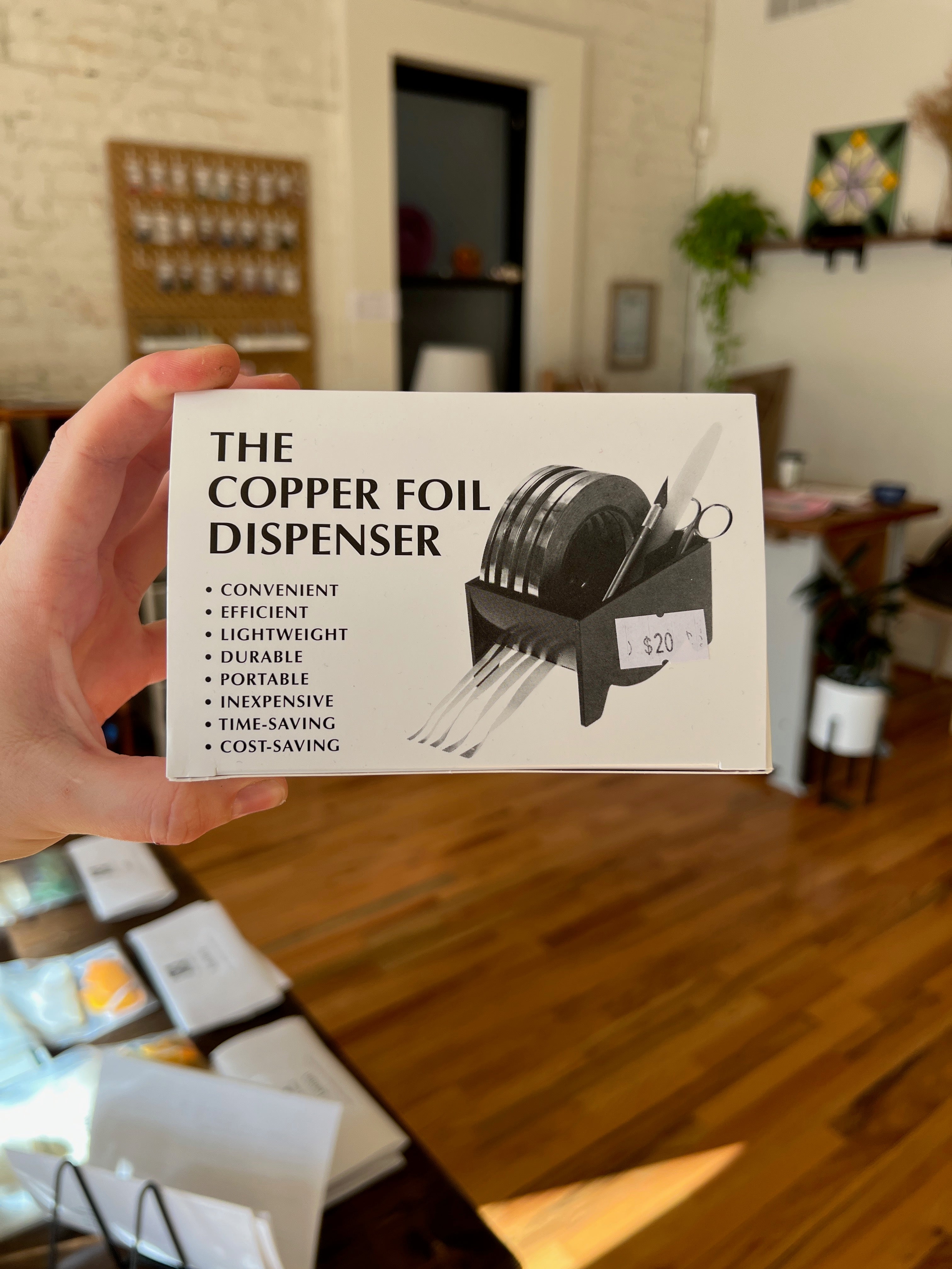 Hand holding a product packaging for 'The Copper Foil Dispenser' with a blurred indoor background.