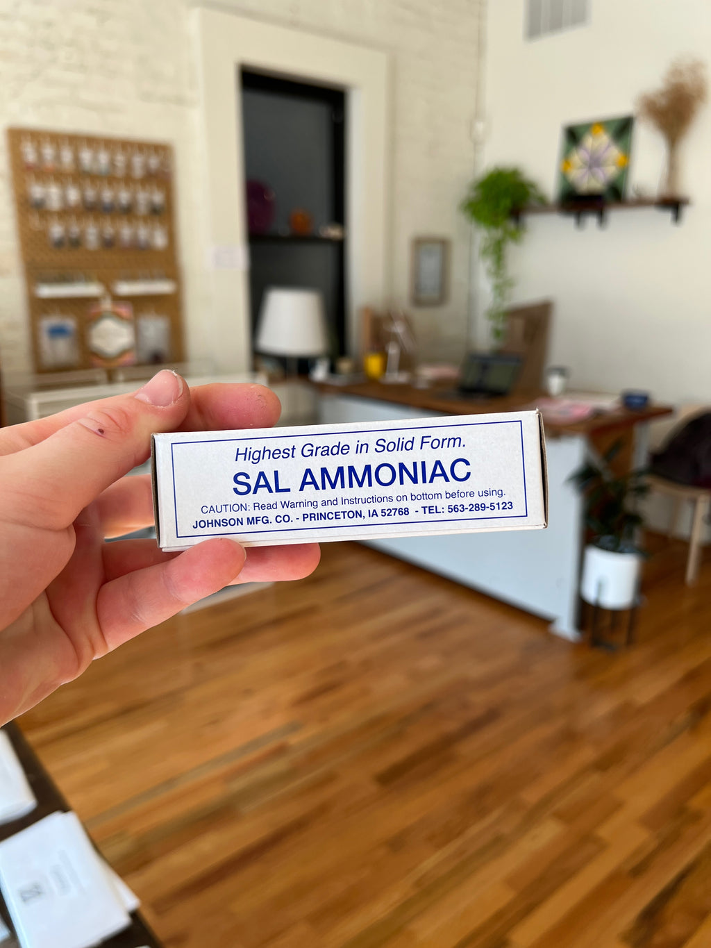Hand holding a label for 'Sal Ammoniac' in a room with wooden floors and furniture.