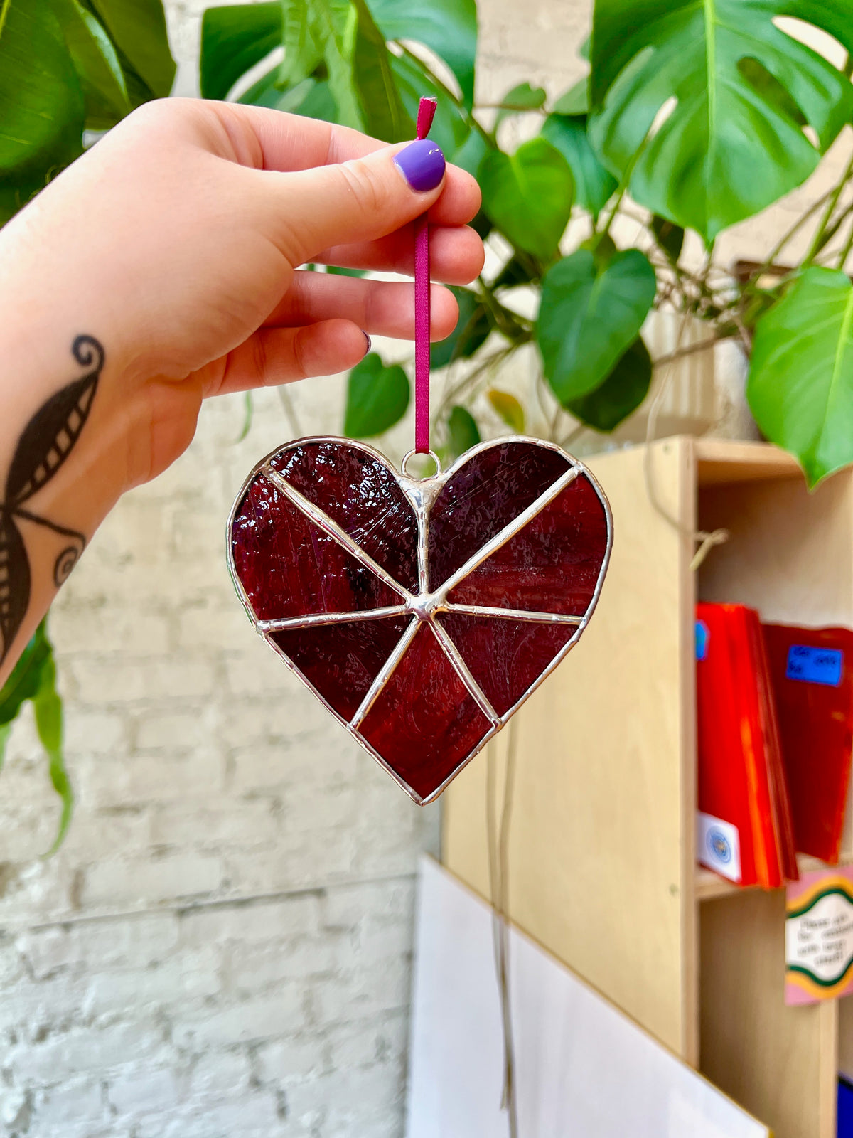 Heart-shaped stained glass ornament held by a hand with purple nail polish, against a blurred indoor background.
