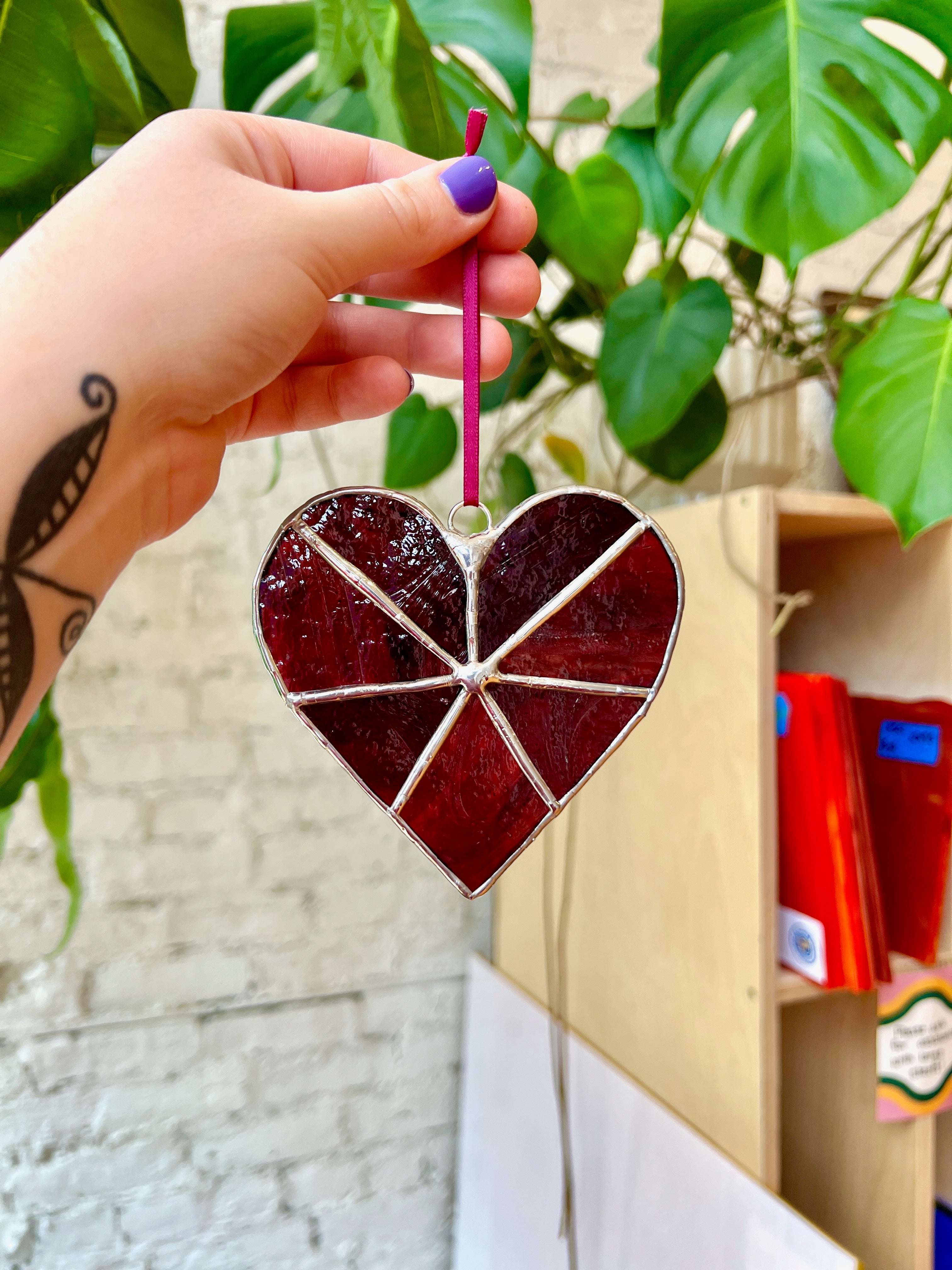Heart-shaped stained glass ornament held by a hand with purple nail polish, against a blurred indoor background.