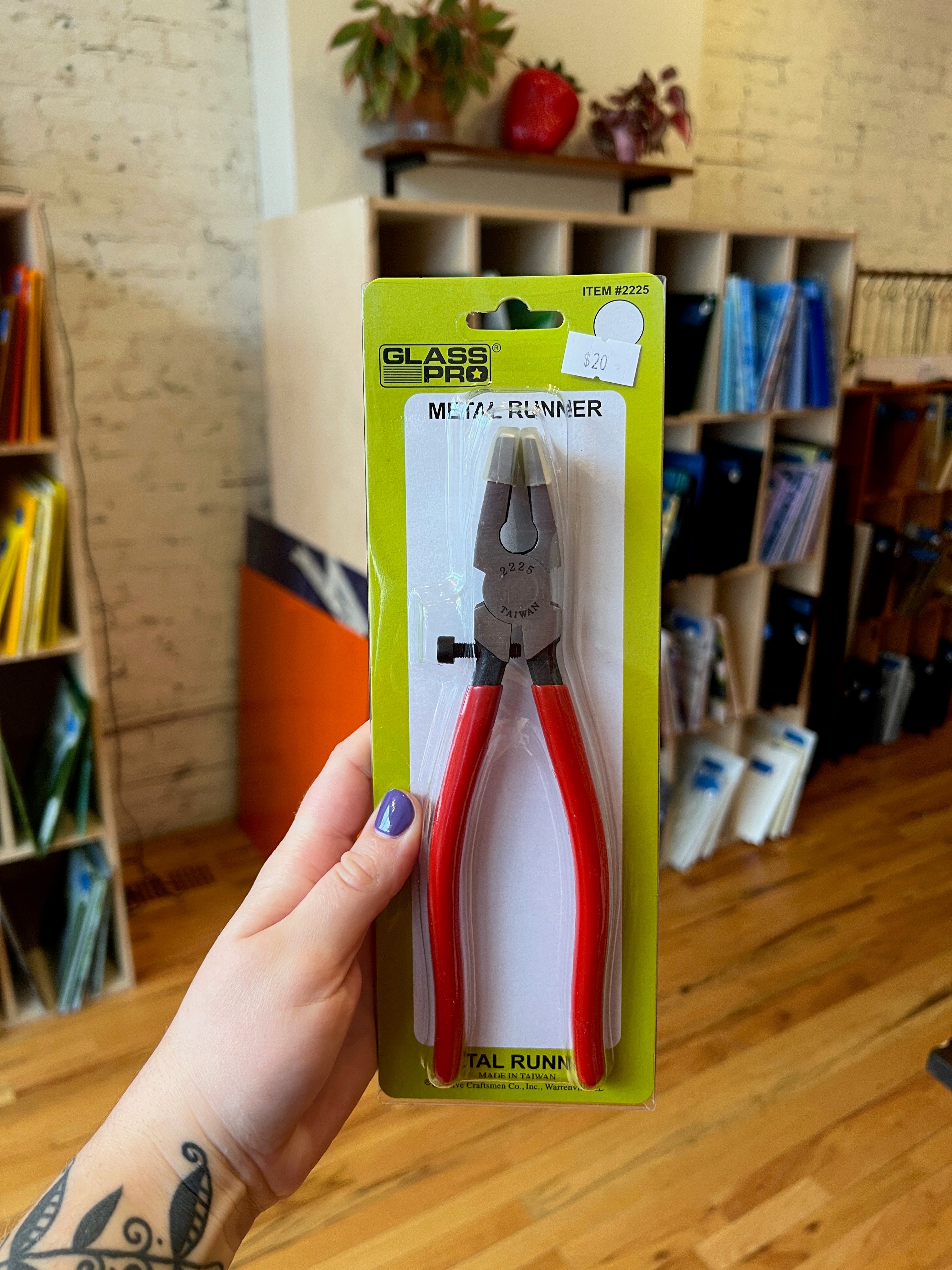 Metal runner pliers in packaging held by a hand with a tattoo, against a blurred indoor background.
