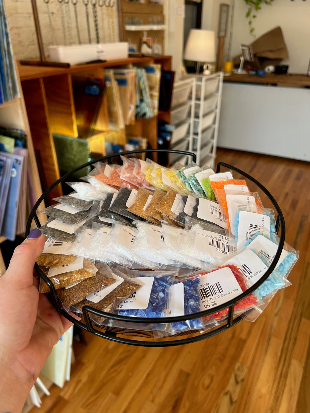 Hand holding a round tray with various small glass frit packages in a store setting 