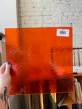 Person holding a textured orange sheet of glass with a barcode label against a brick wall.