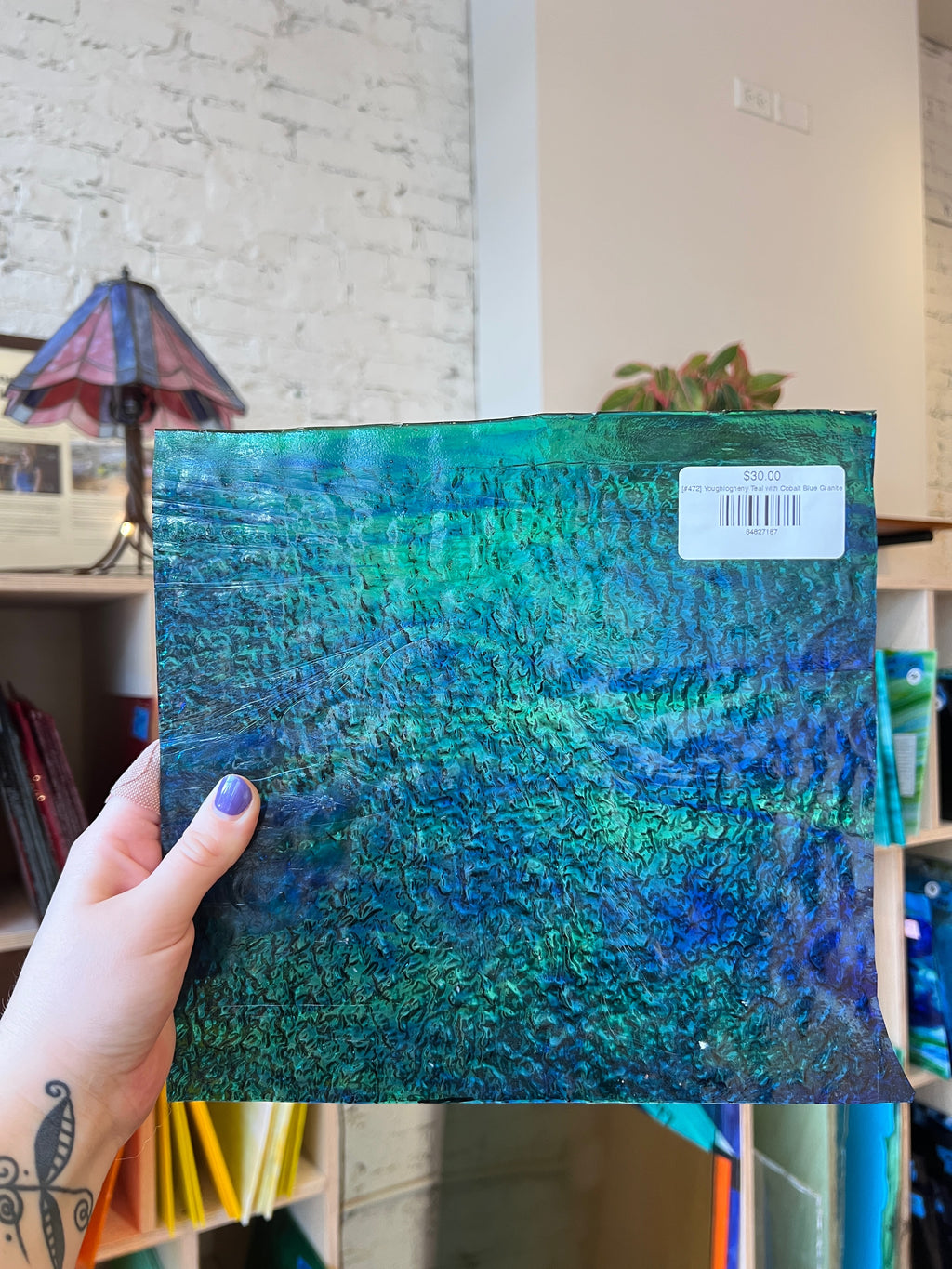 Hand holding a blue and green textured sheet glass with a visible brand label against an interior background.