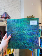Hand holding a blue and green textured sheet glass with a visible brand label against an interior background.
