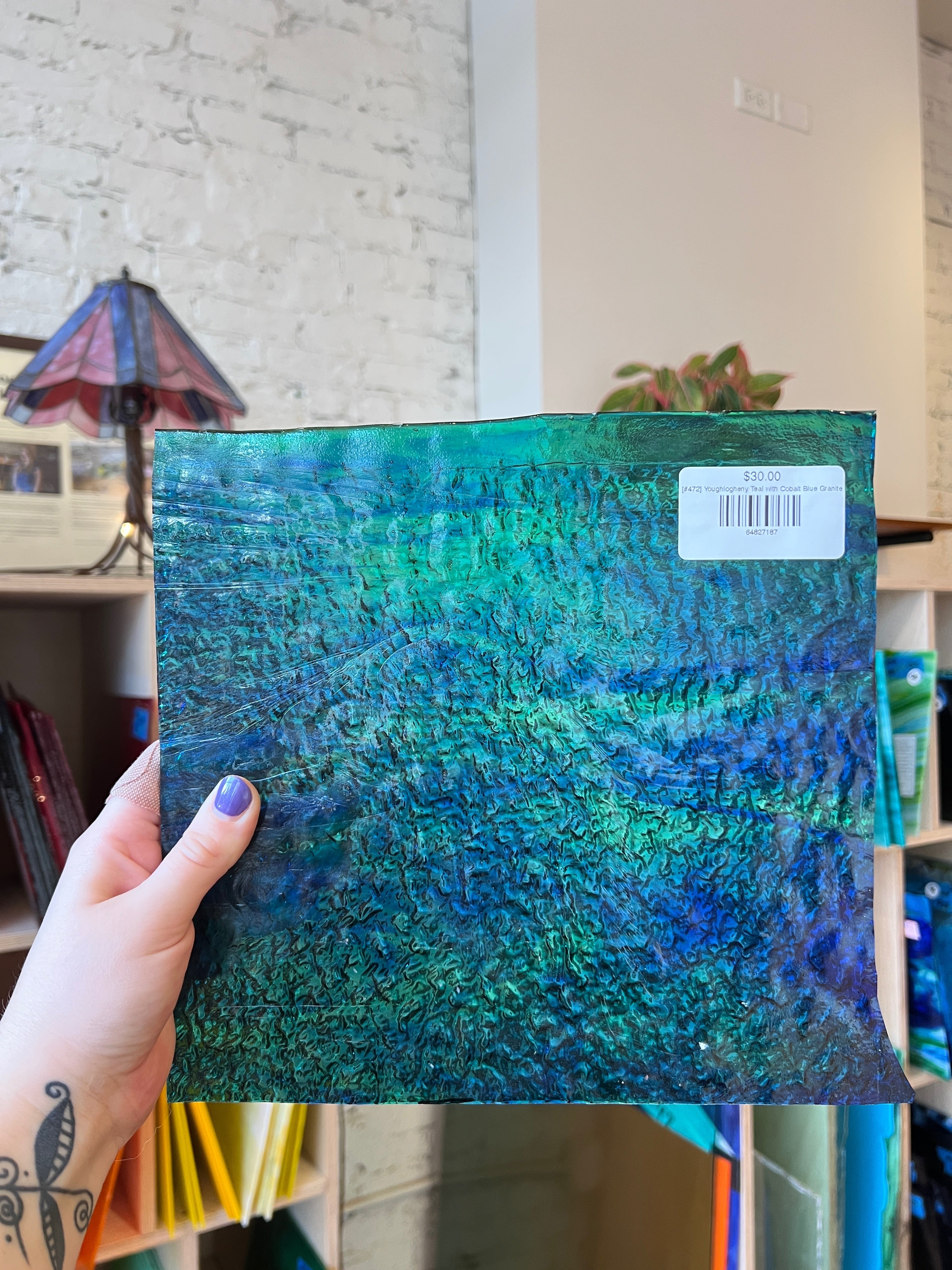 Hand holding a blue and green textured sheet glass with a visible brand label against an interior background.