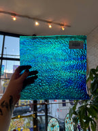 Hand holding a textured blue and green sheet glass panel with a blurred indoor background
