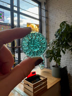 Person holding a green circular glass gem with intricate patterns in front of a window.