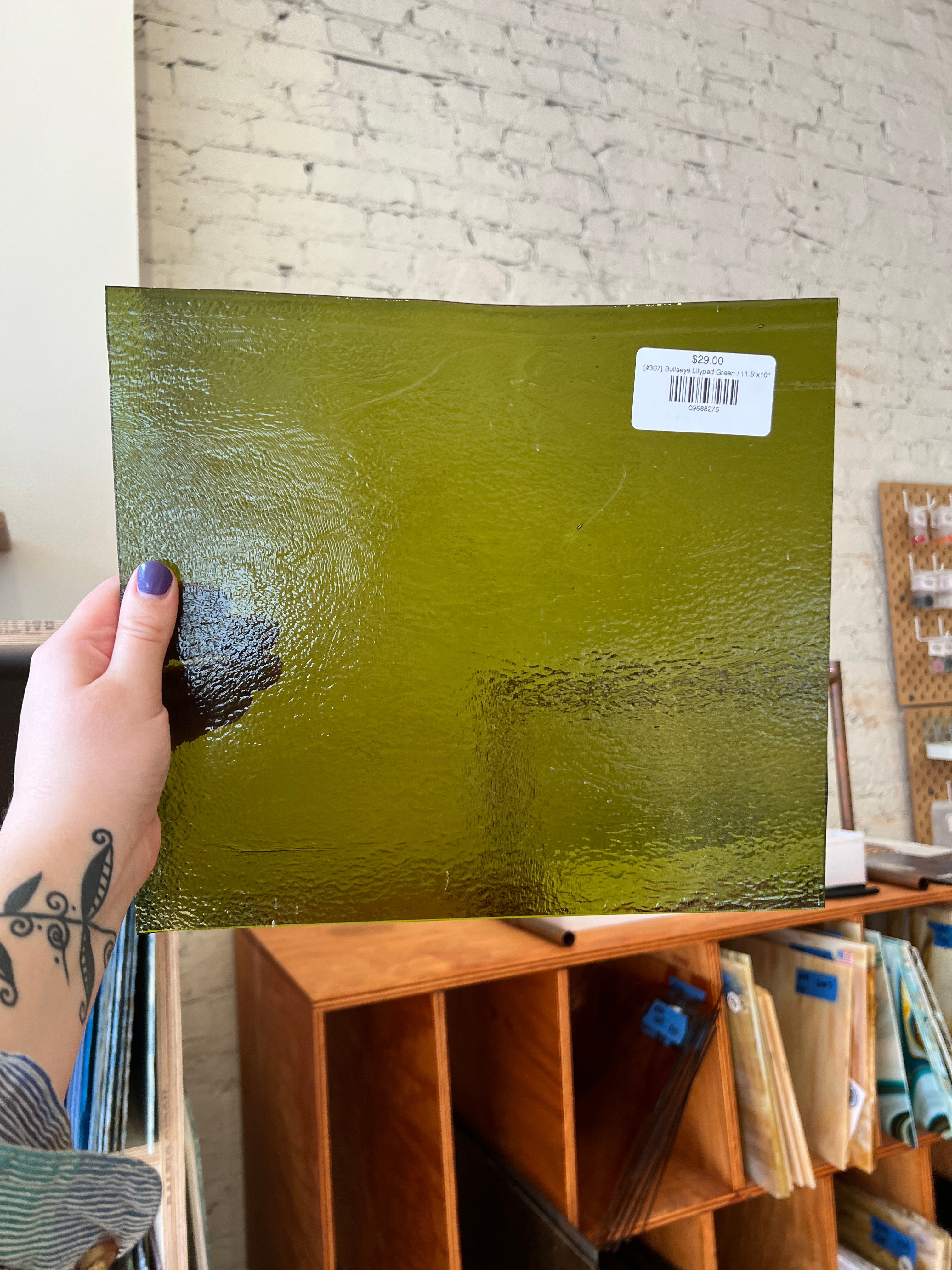 Green textured glass sheet held by a hand with a tattoo, against a background of wooden shelves.