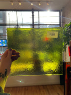 Green textured glass panel held by a hand with a tattoo, indoors.