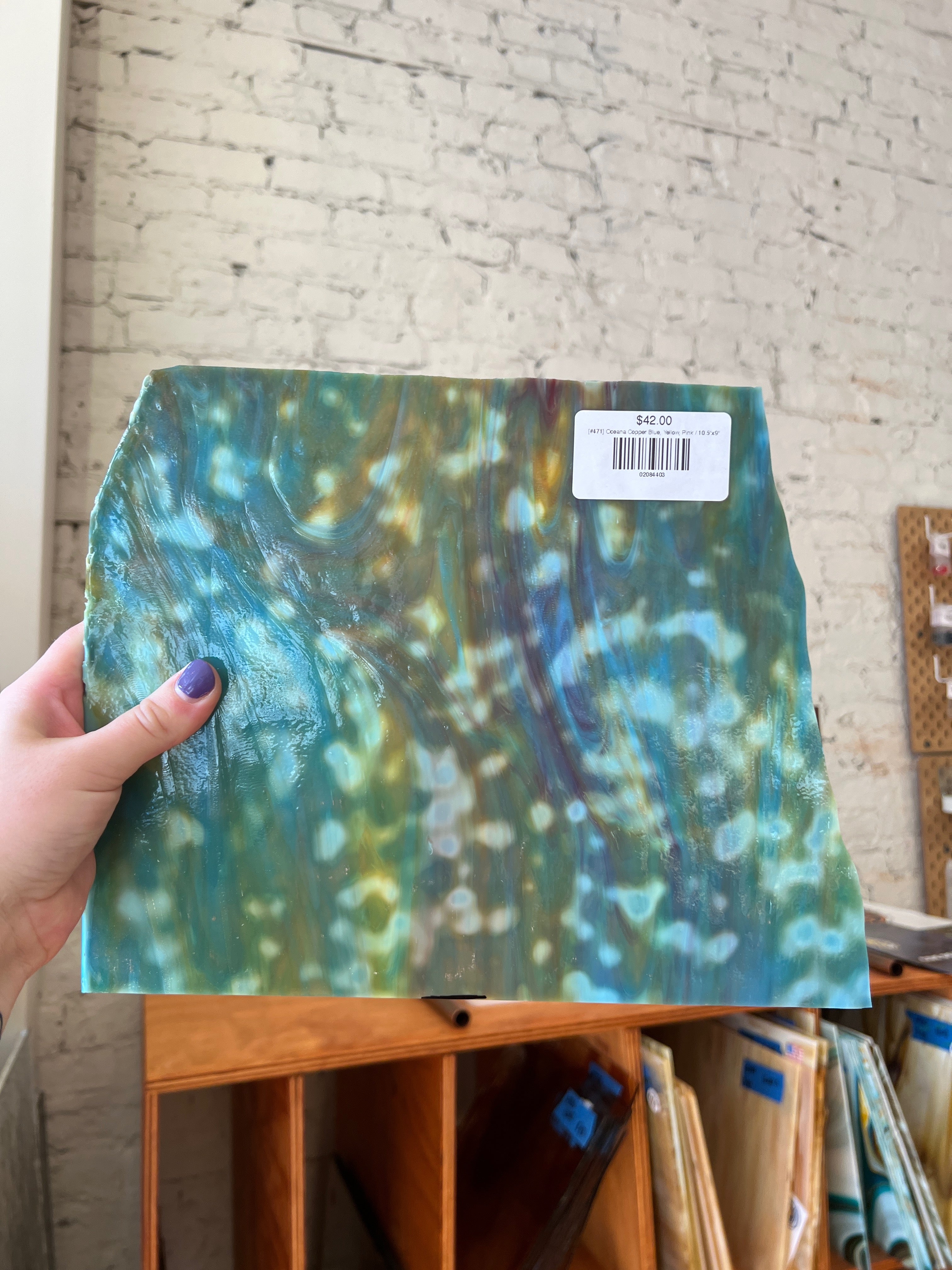 Hand holding a piece of sheet glass with a colorful pattern against a brick wall.