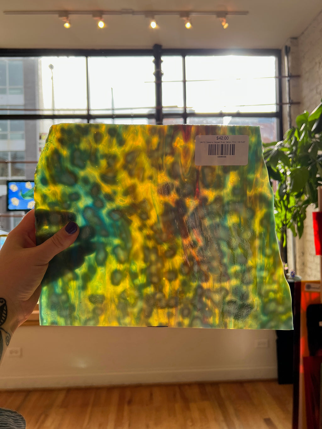 Colorful tie-dye patterned sheet glass held by a hand indoors with large windows.