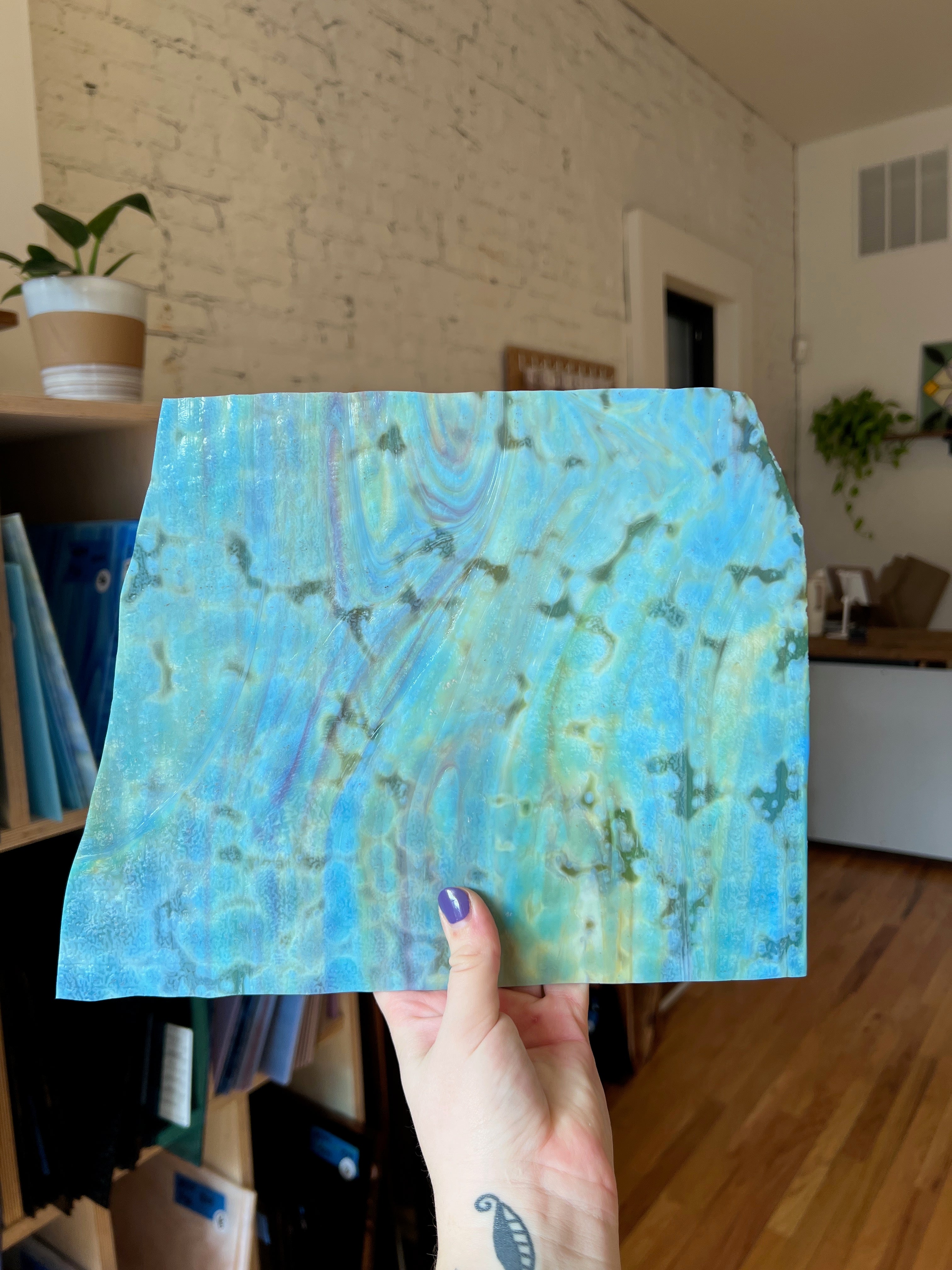 Hand holding a piece of sheet glass with blue and green abstract design in a room.