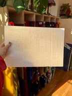 Person holding a large white sheet of glass in front of a bookshelf with various items.