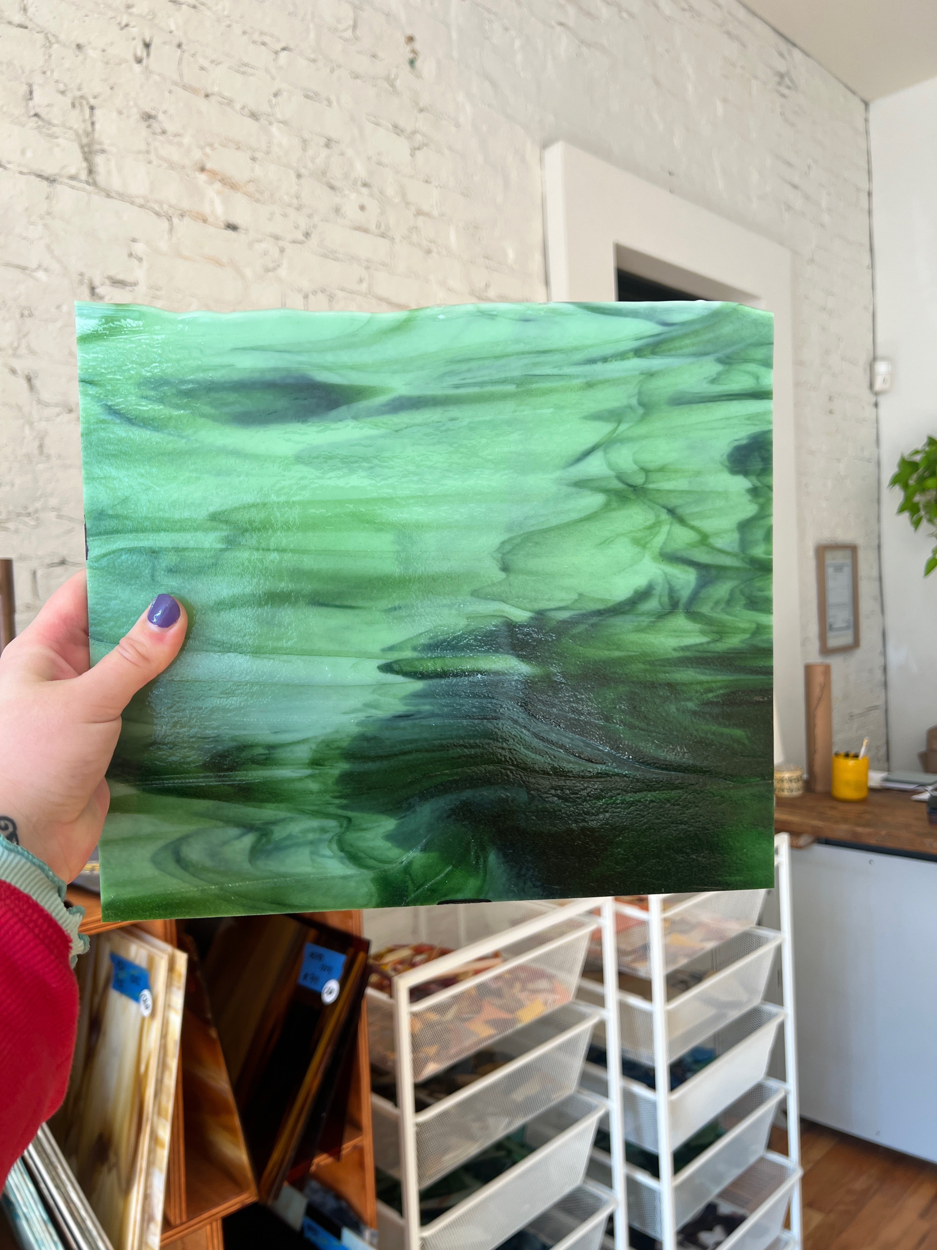 Person holding a green marbled piece of stained glass against a white brick wall.