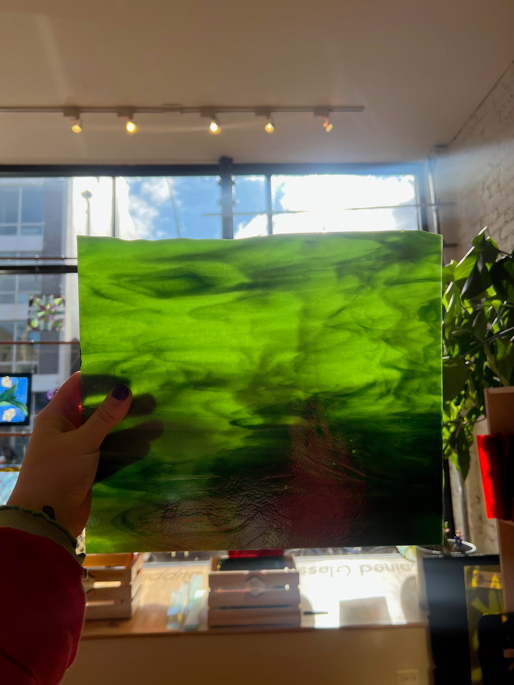 Person holding a large green piece of stained glass indoors with a window in the background