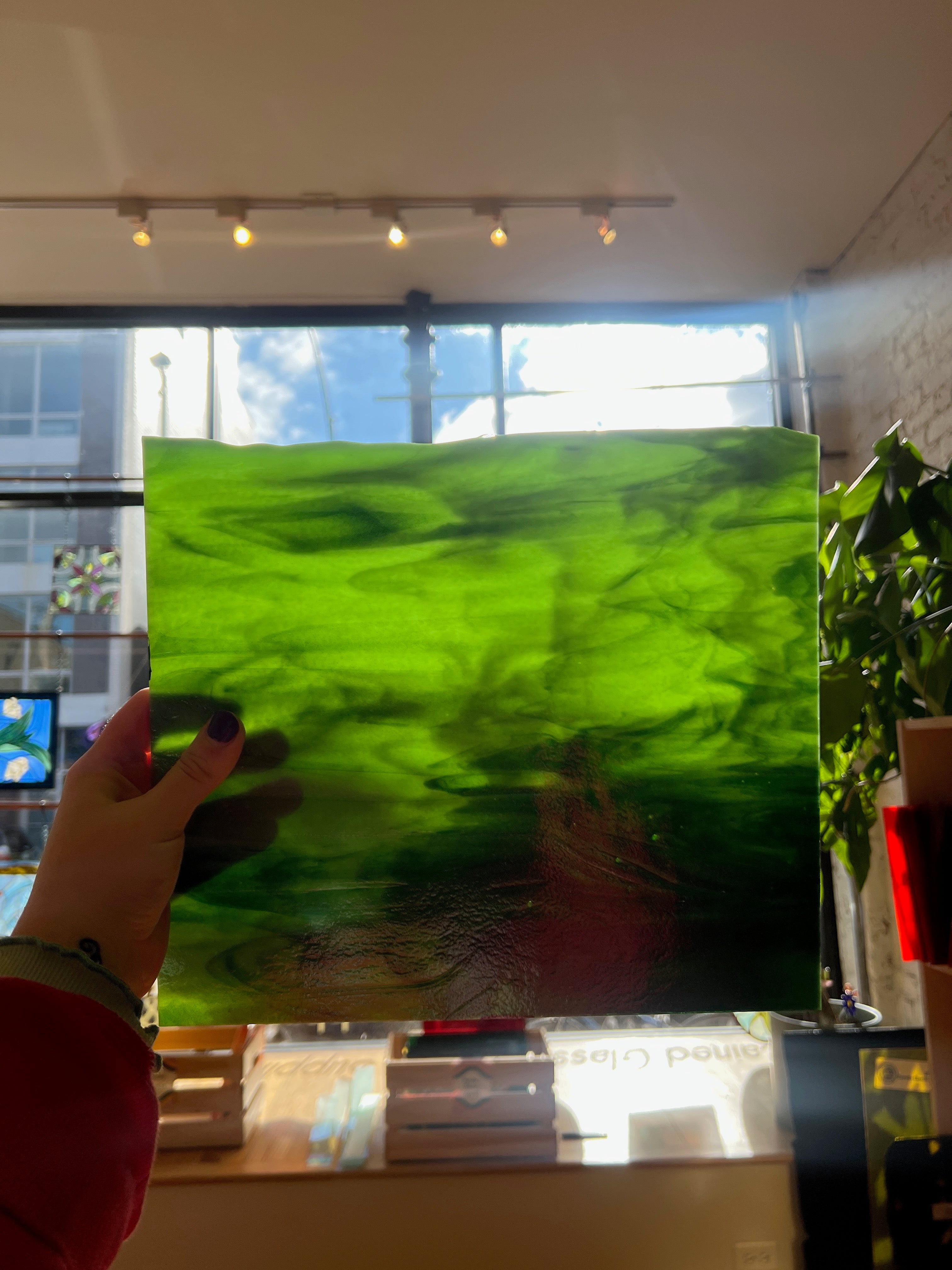 Person holding a large green piece of stained glass indoors with a window in the background