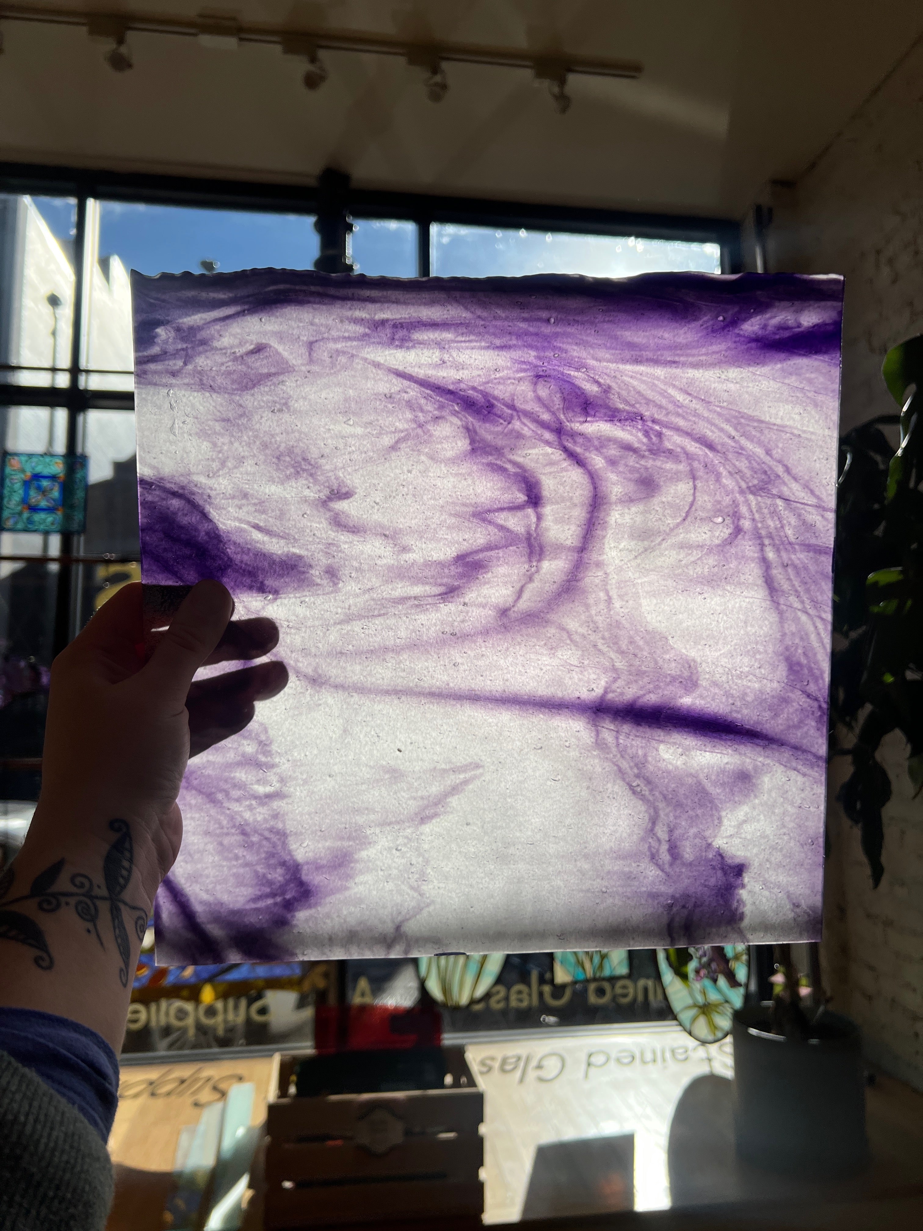 Hand holding a piece of glass with purple and white abstract pattern in front of a window.