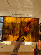 Hand holding a large swirled piece of glass with warm colors in an indoor setting.