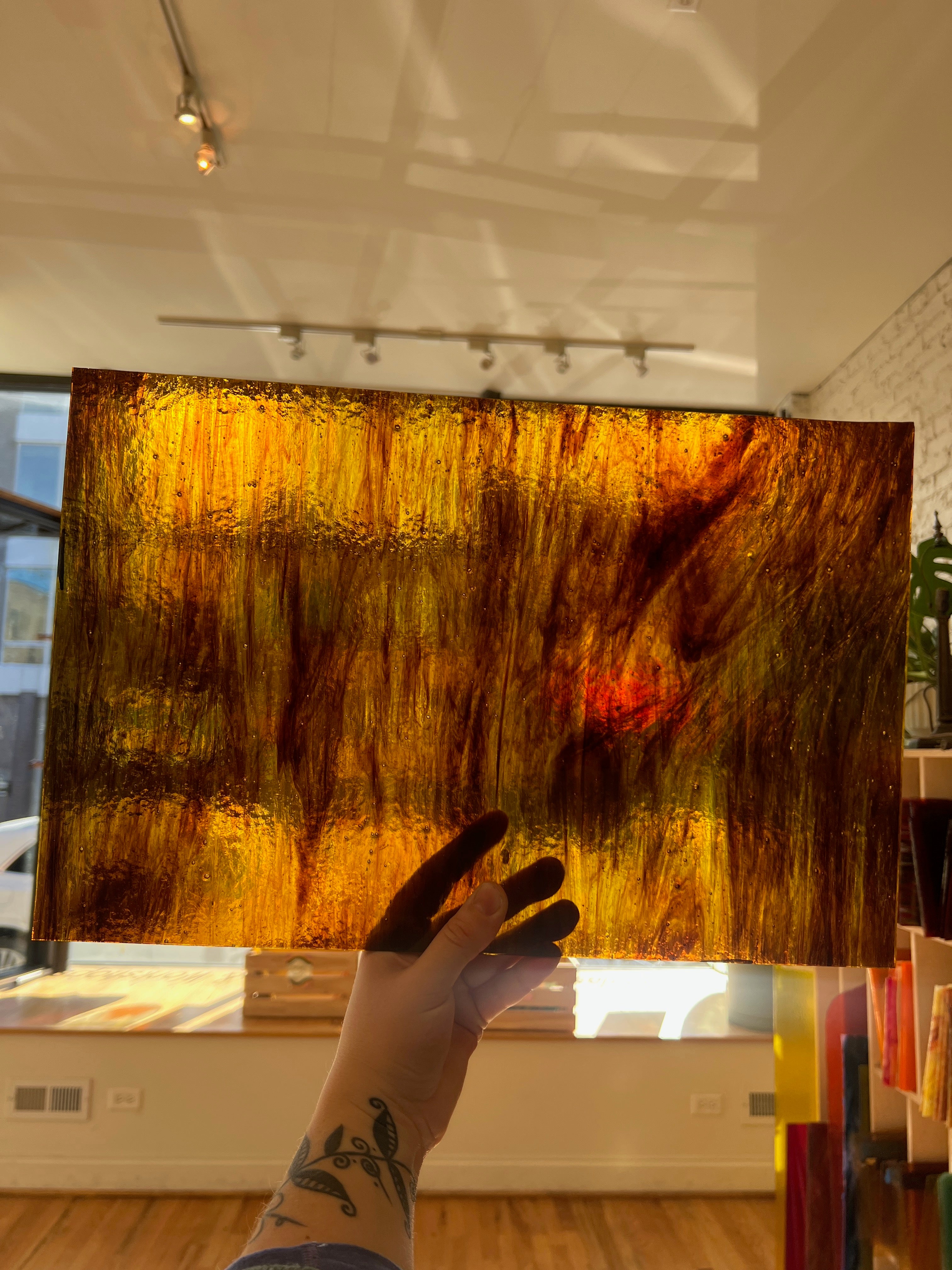 Hand holding a large swirled piece of glass with warm colors in an indoor setting.