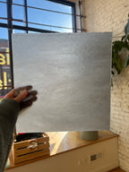 Person holding a large gray piece of stained glass against a window with a plant above