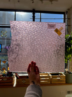 Hand holding a textured pink sheet of glass in front of a window with a plant and decorative items in the background.