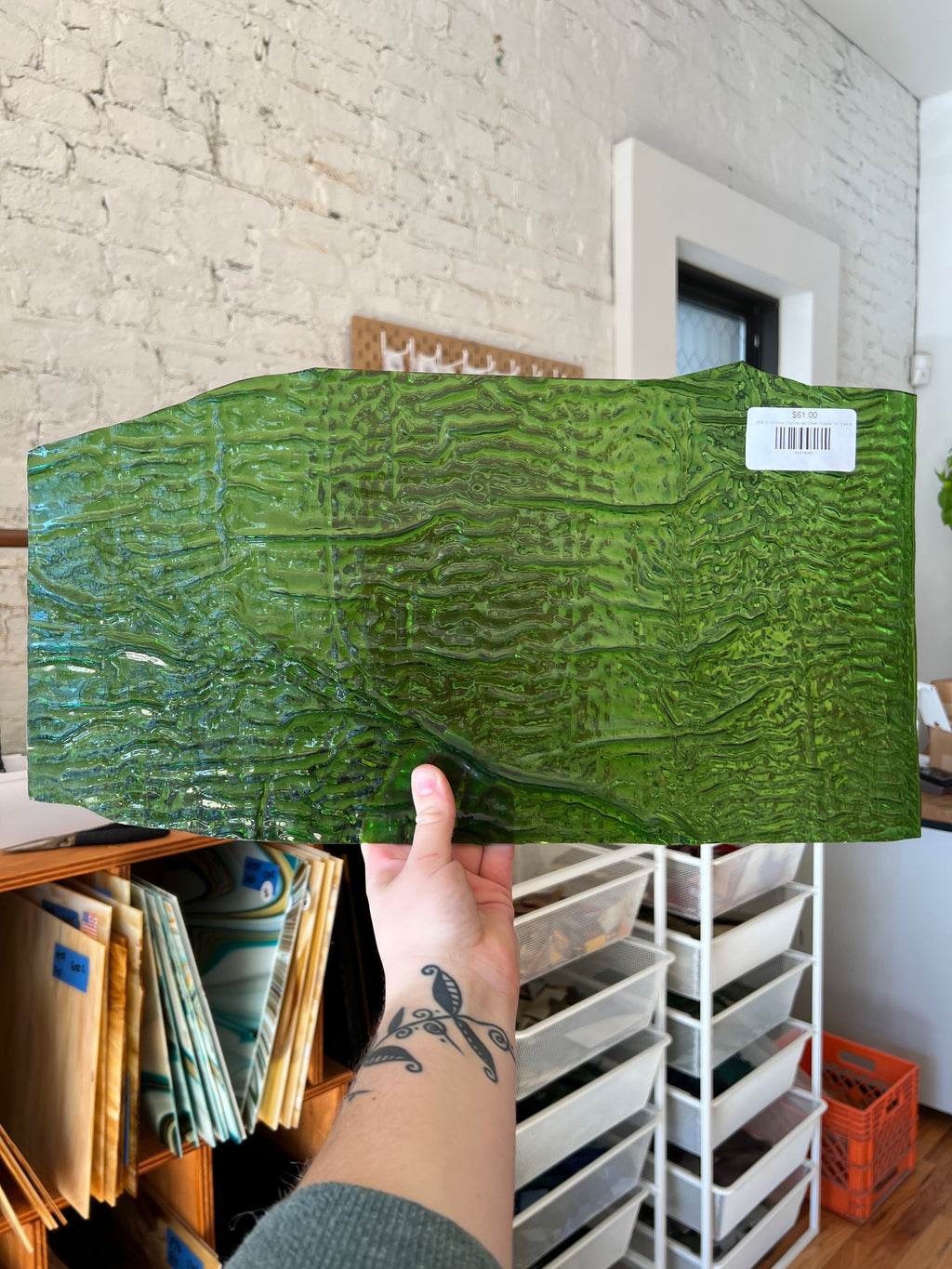 Person holding a large green textured stained glass sheet in a room with white brick walls and storage shelves.