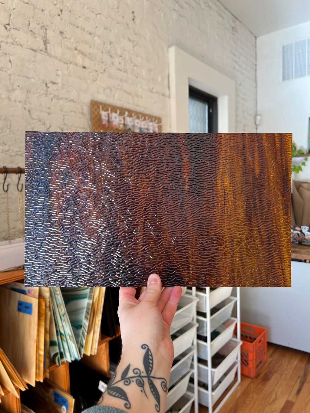 Person holding a textured brown piece of stained glass in a room with white brick walls and wooden floors.
