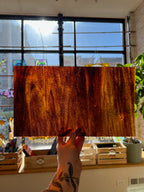 Hand holding a textured brown rectangular piece of stained glass in front of a window with a view of trees.