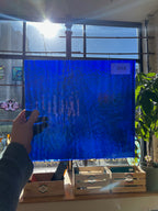 Person holding a blue textured piece of stained glass in front of a window