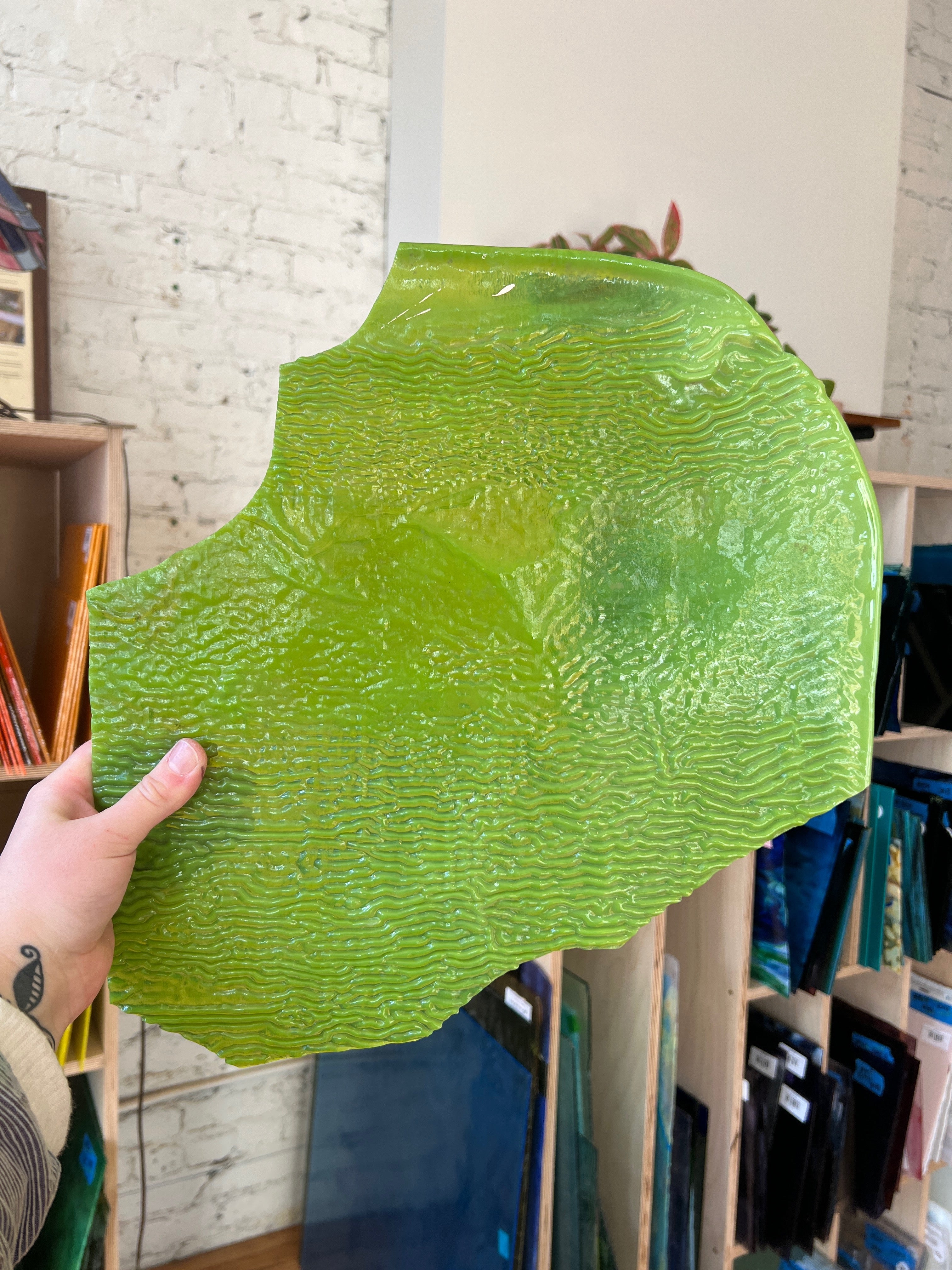 Person holding a large green textured piece of stained glass indoors with a bookshelf in the background
