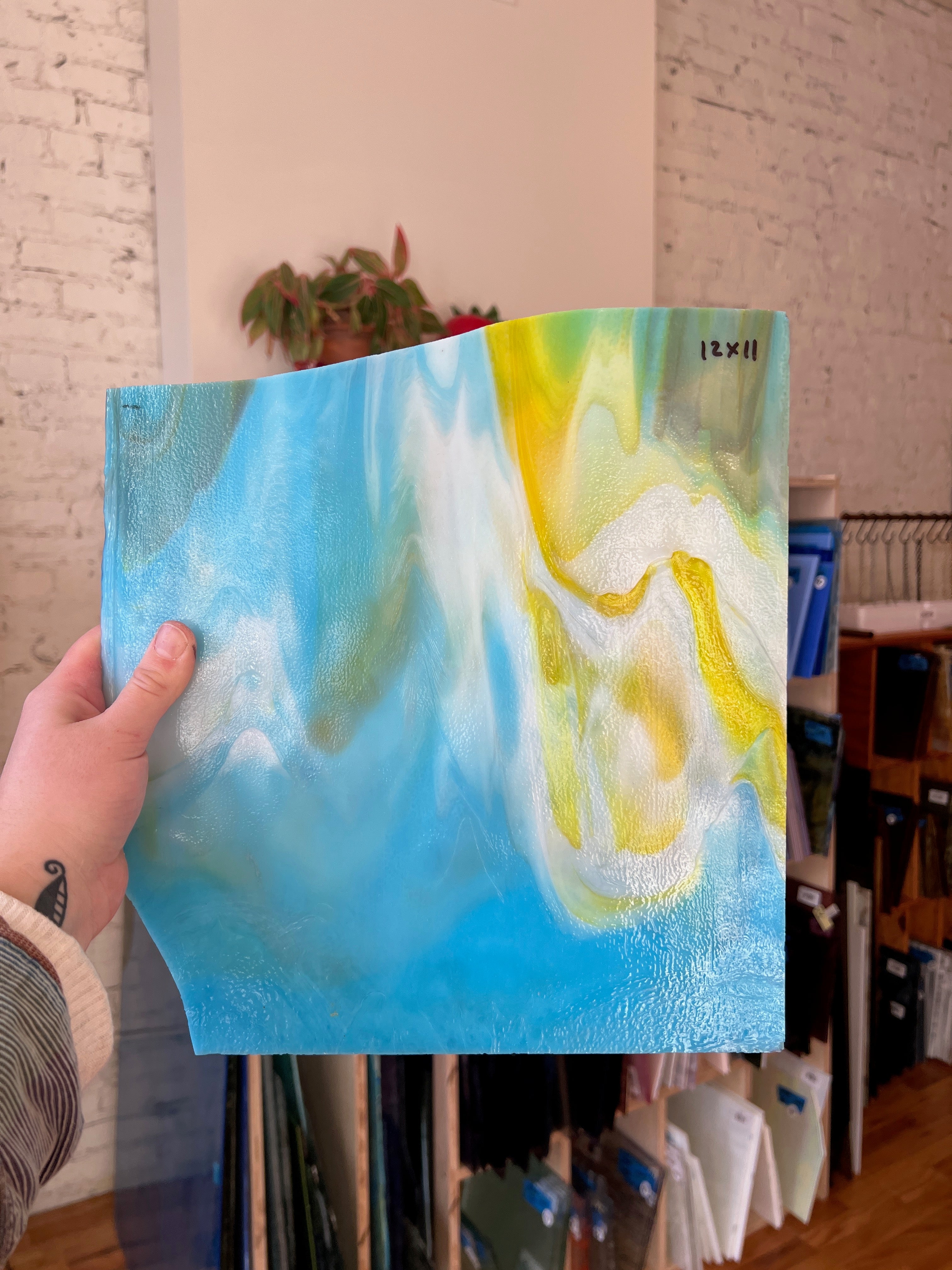 Person holding a piece of blue and yellow swirled stained glass in front of a blurred interior.