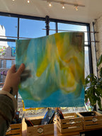 Person holding a piece of blue and yellow swirled stained glass in front of a blurred window.