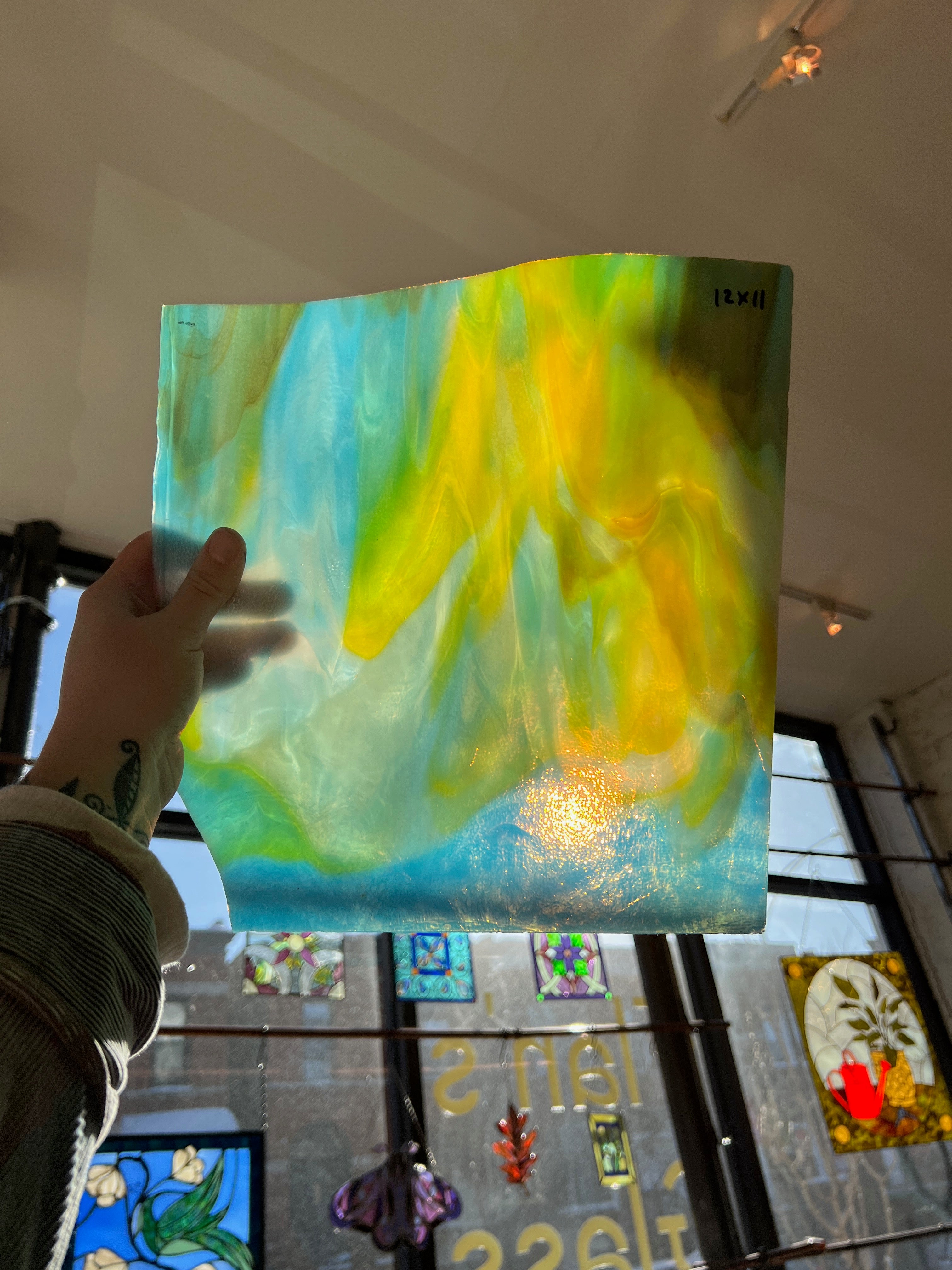 Person holding a piece of blue and yellow swirled stained glass in front of a blurred window.