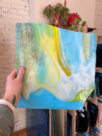 Person holding a piece of blue and yellow swirled stained glass in front of a blurred interior.