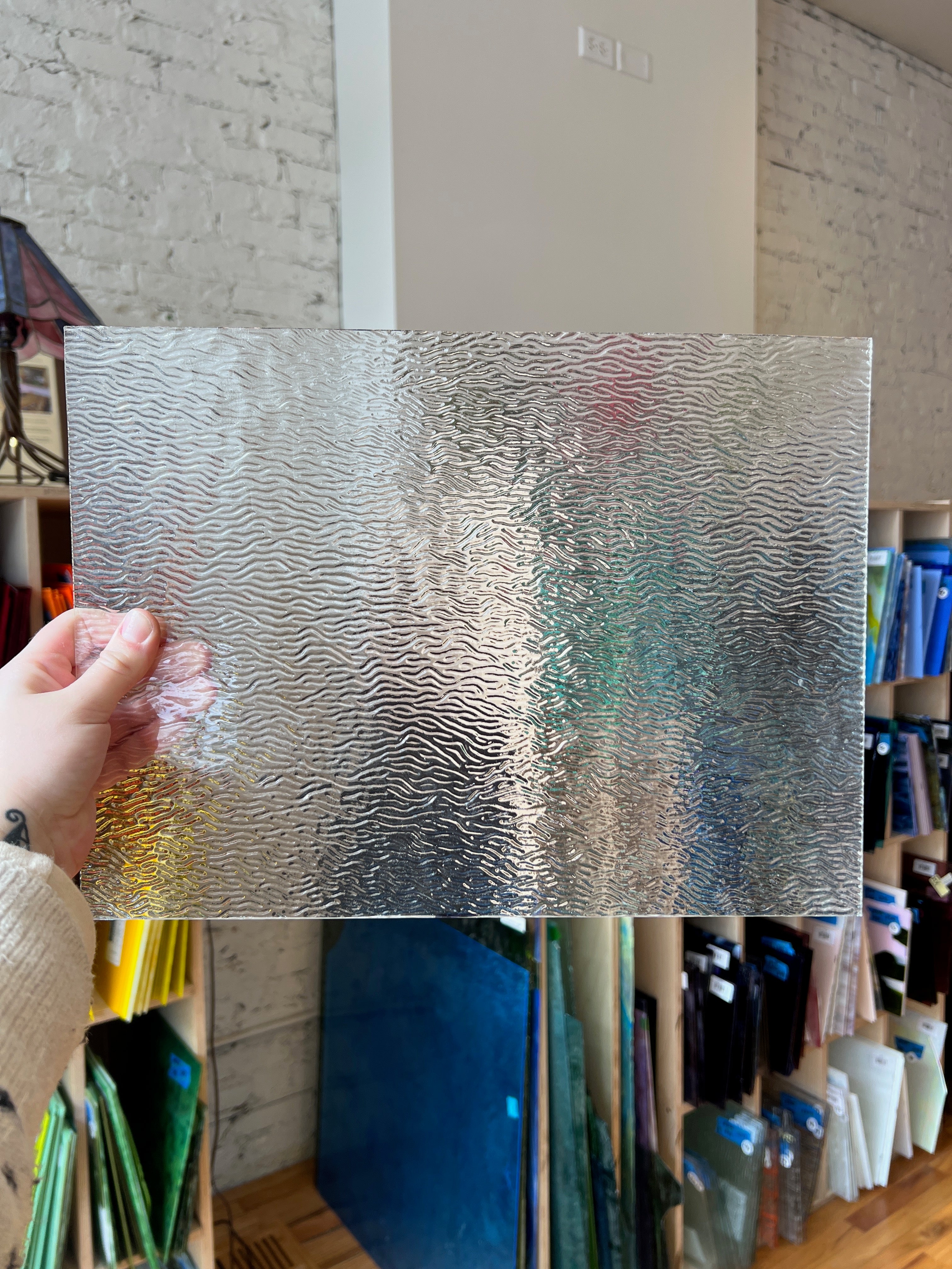 Hand holding a sheet of textured glass against a background with shelves.