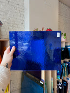 Person holding a piece of blue textured stained glass in front of a blurred interior.