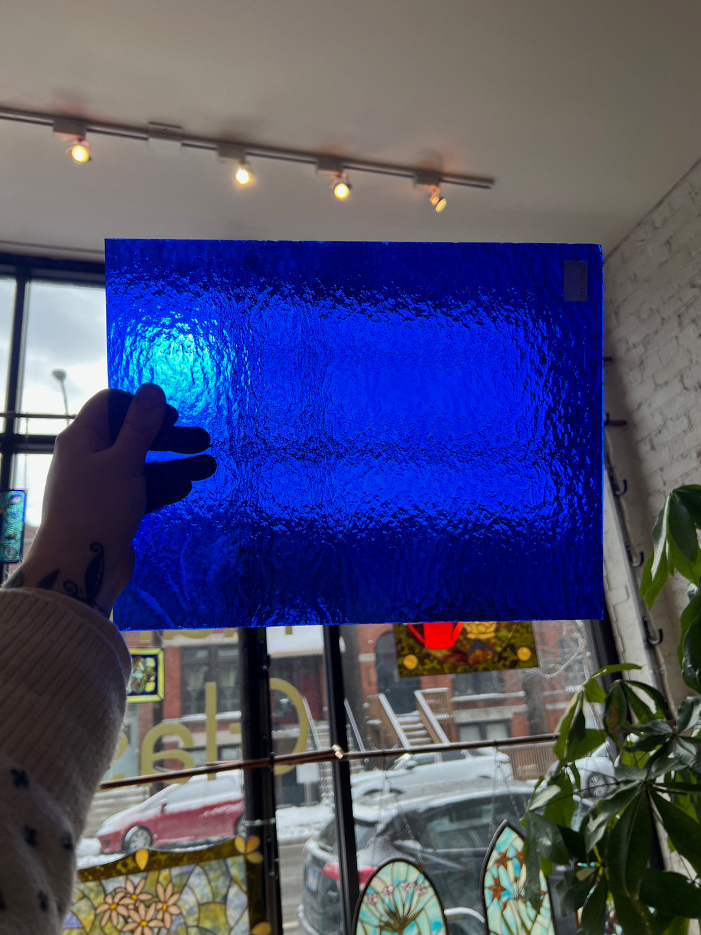 Person holding a piece of blue textured stained glass in front of a blurred window.