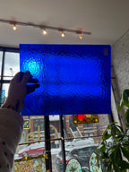 Person holding a piece of blue textured stained glass in front of a blurred window.
