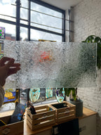 Person holding a frosted textured glass panel in front of a window with stained glass and a white brick wall.