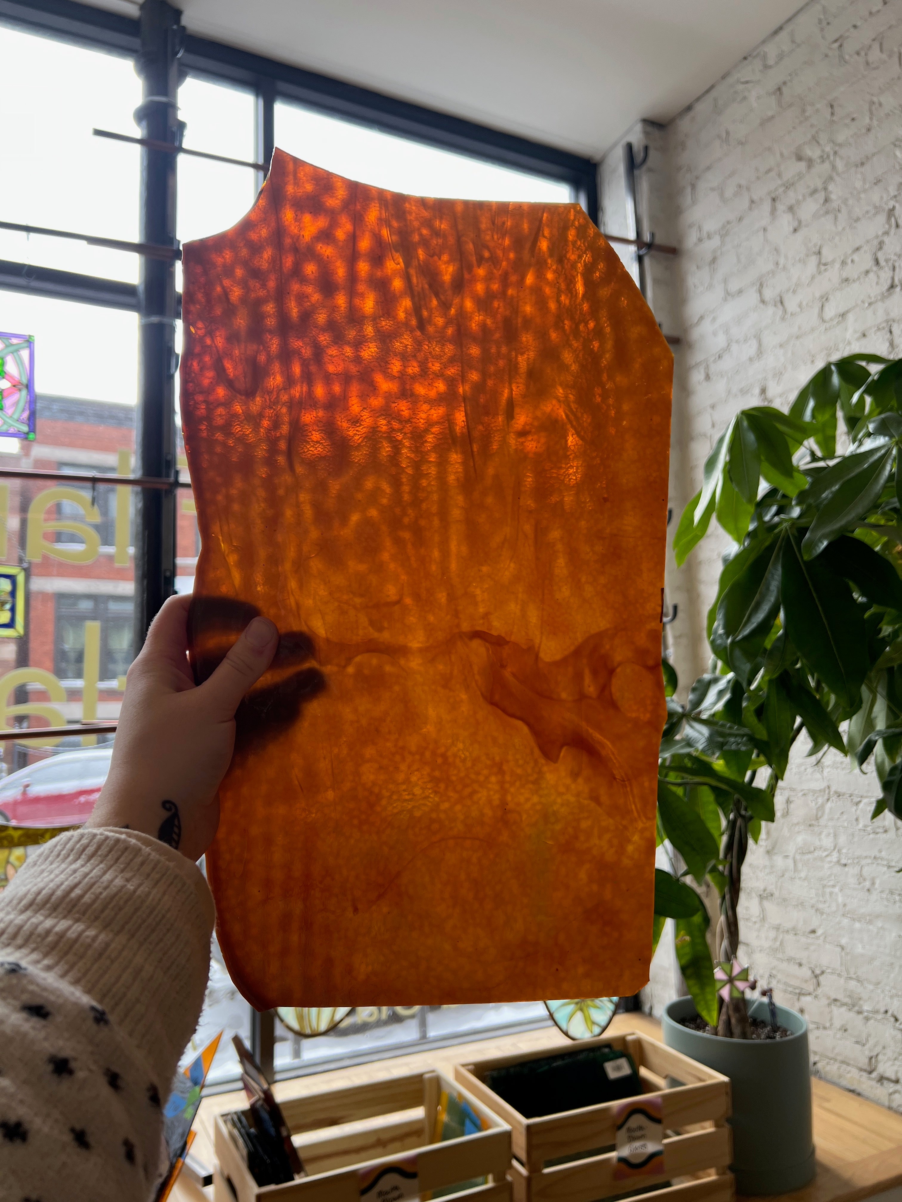 Person holding a piece of amber stained glass in front of a blurred window.