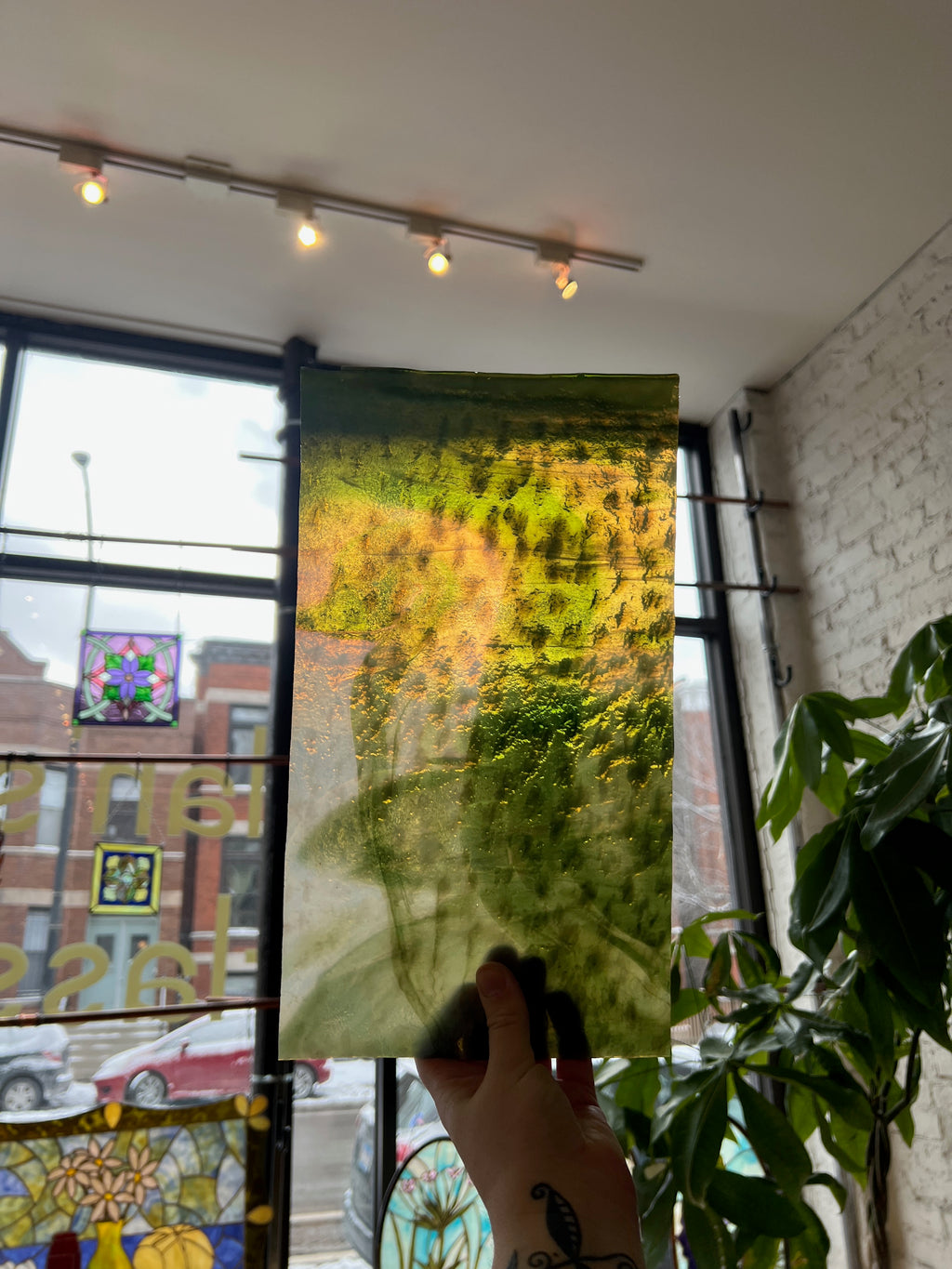 Person holding a piece of green and white stained glass in front of a blurred window.