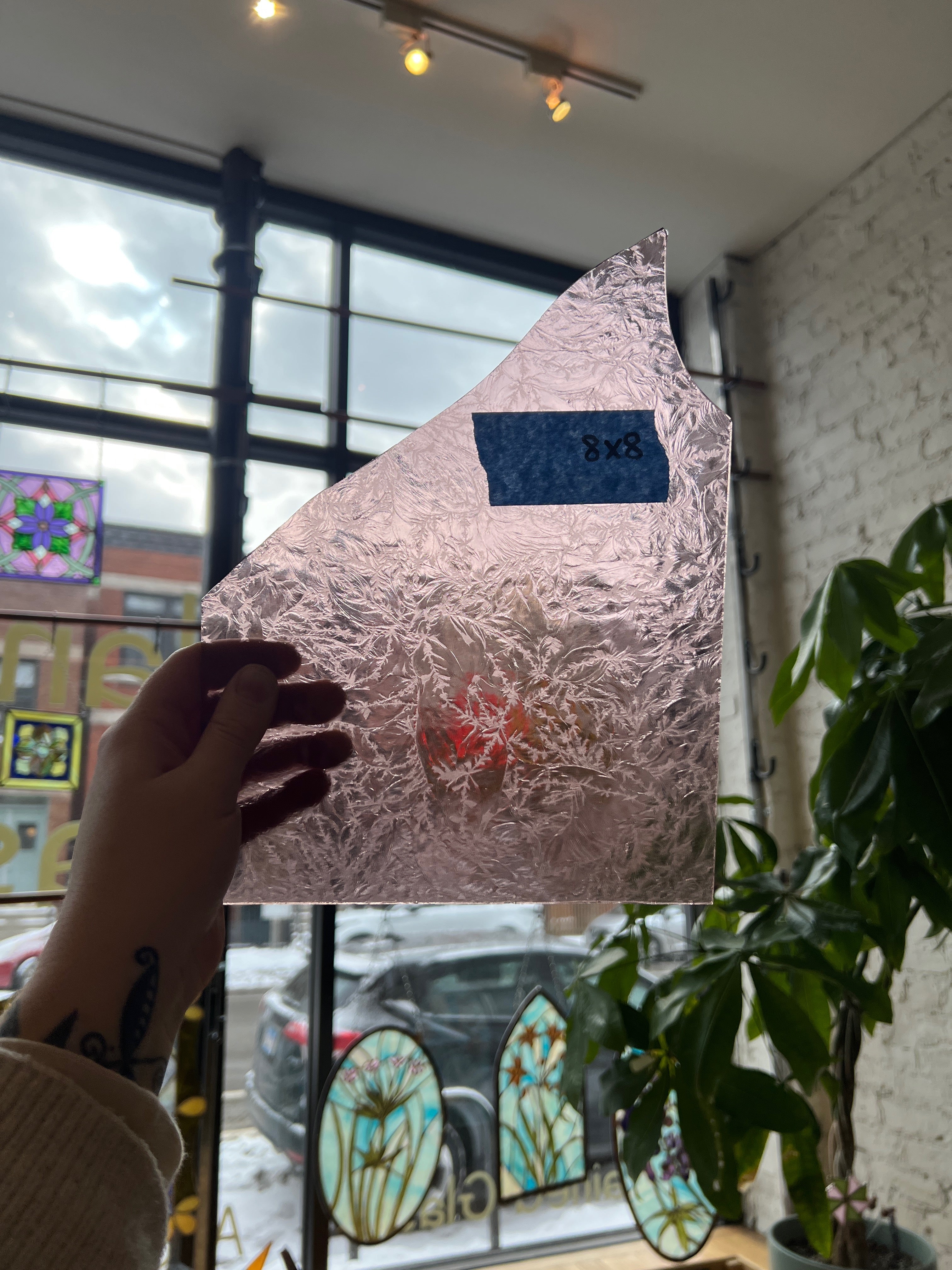 Person holding a lilac transparent textured piece of stained glass in front of a blurred sunny window. 
